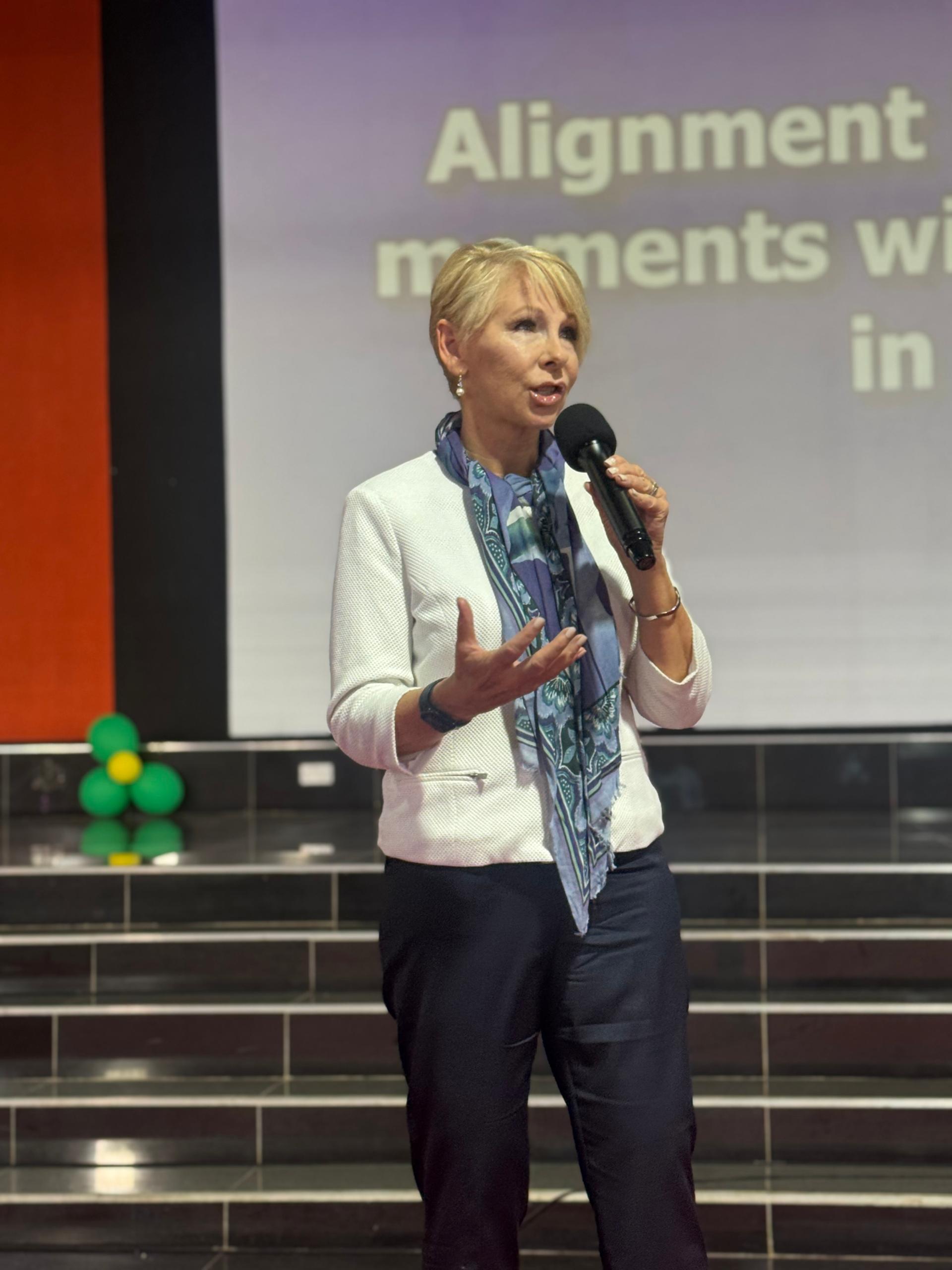 Woman with short blonde hair speaking into a microphone on stage, gesturing with her hand, with a presentation slide behind her.