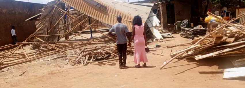 Two people stand in front of a collapsed wooden structure with scattered debris around them.