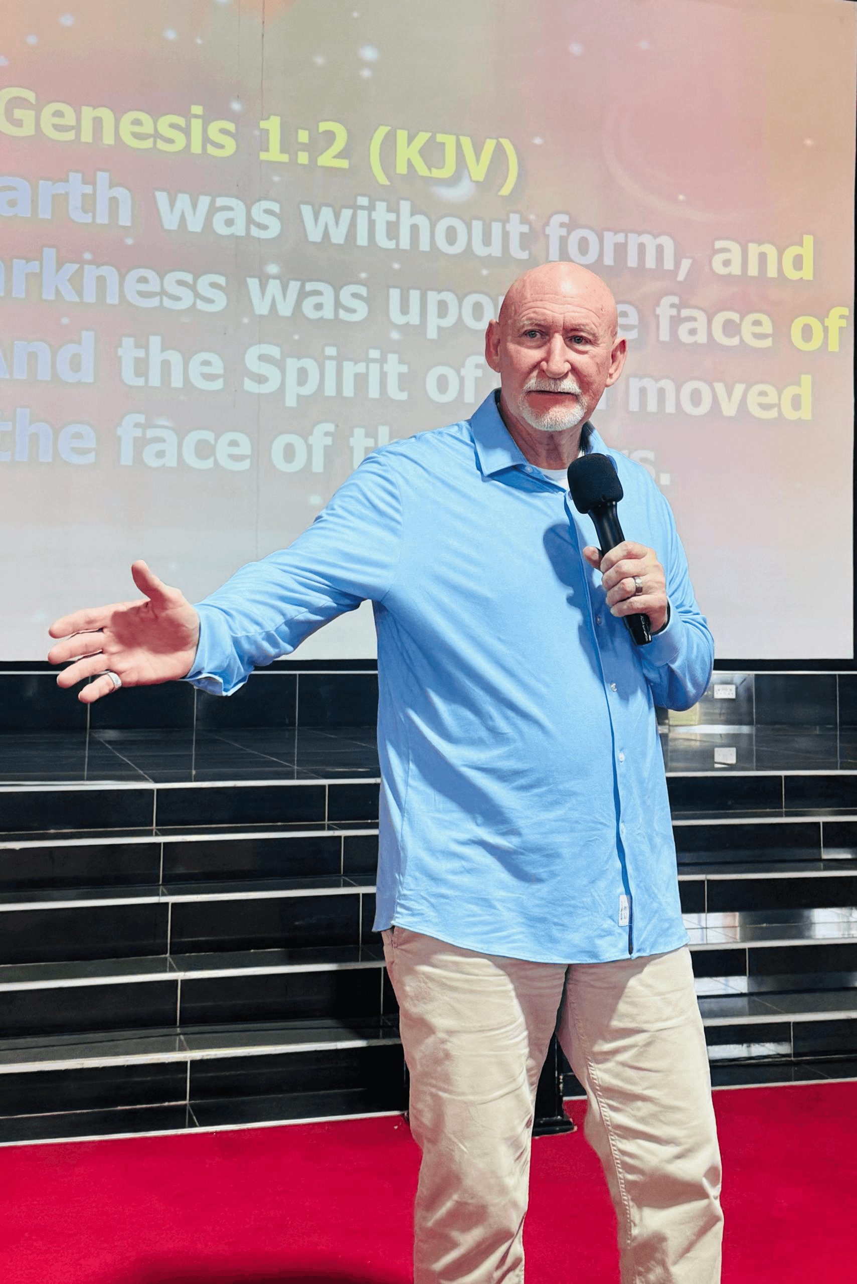 Older man with white hair and beard speaking into a microphone on stage, gesturing with his left hand, in front of a screen.