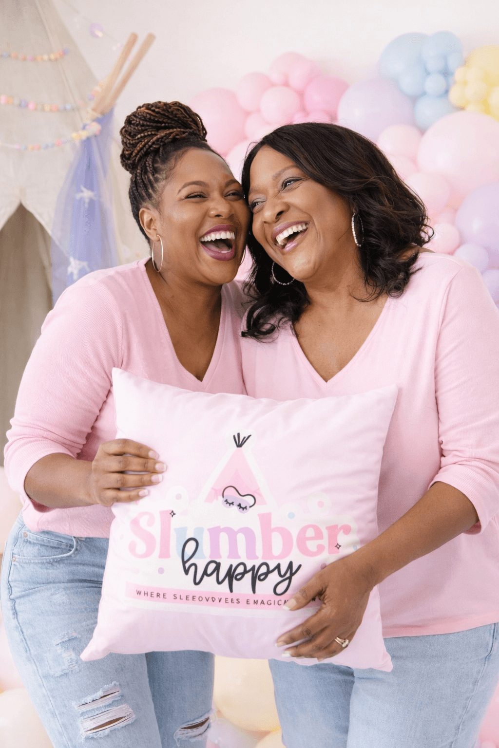 Two women smiling and holding a pillow with birthday message, surrounded by pastel balloons.
