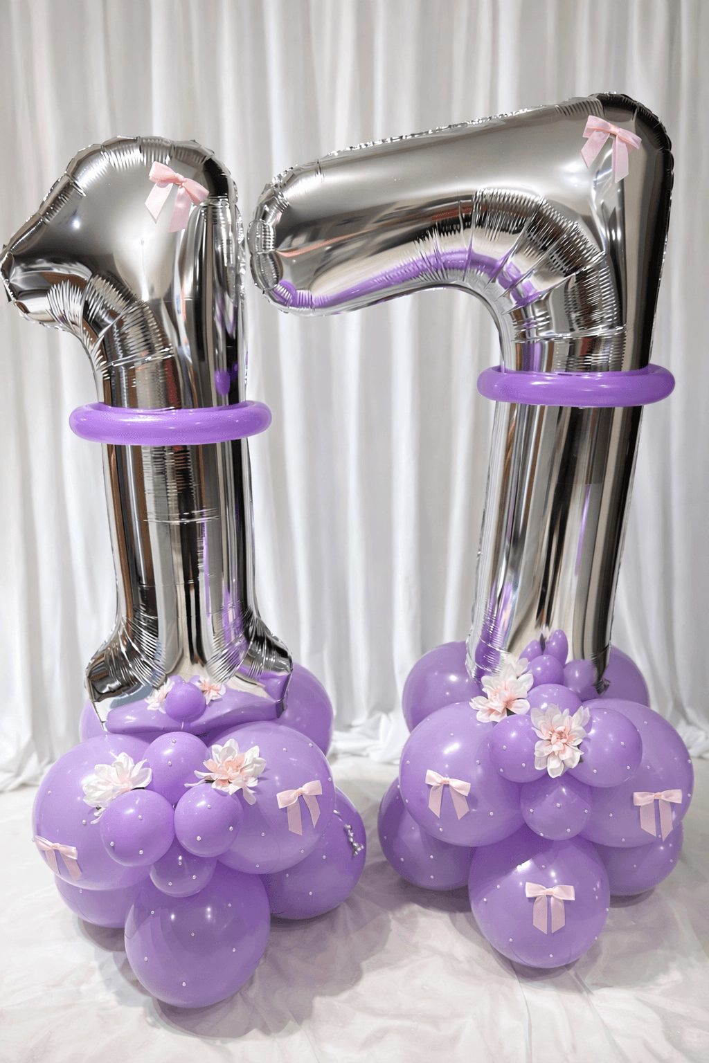 Two large silver number balloons '1' and '7' with purple balloon bases decorated with flowers and crosses, set against a white curtain.