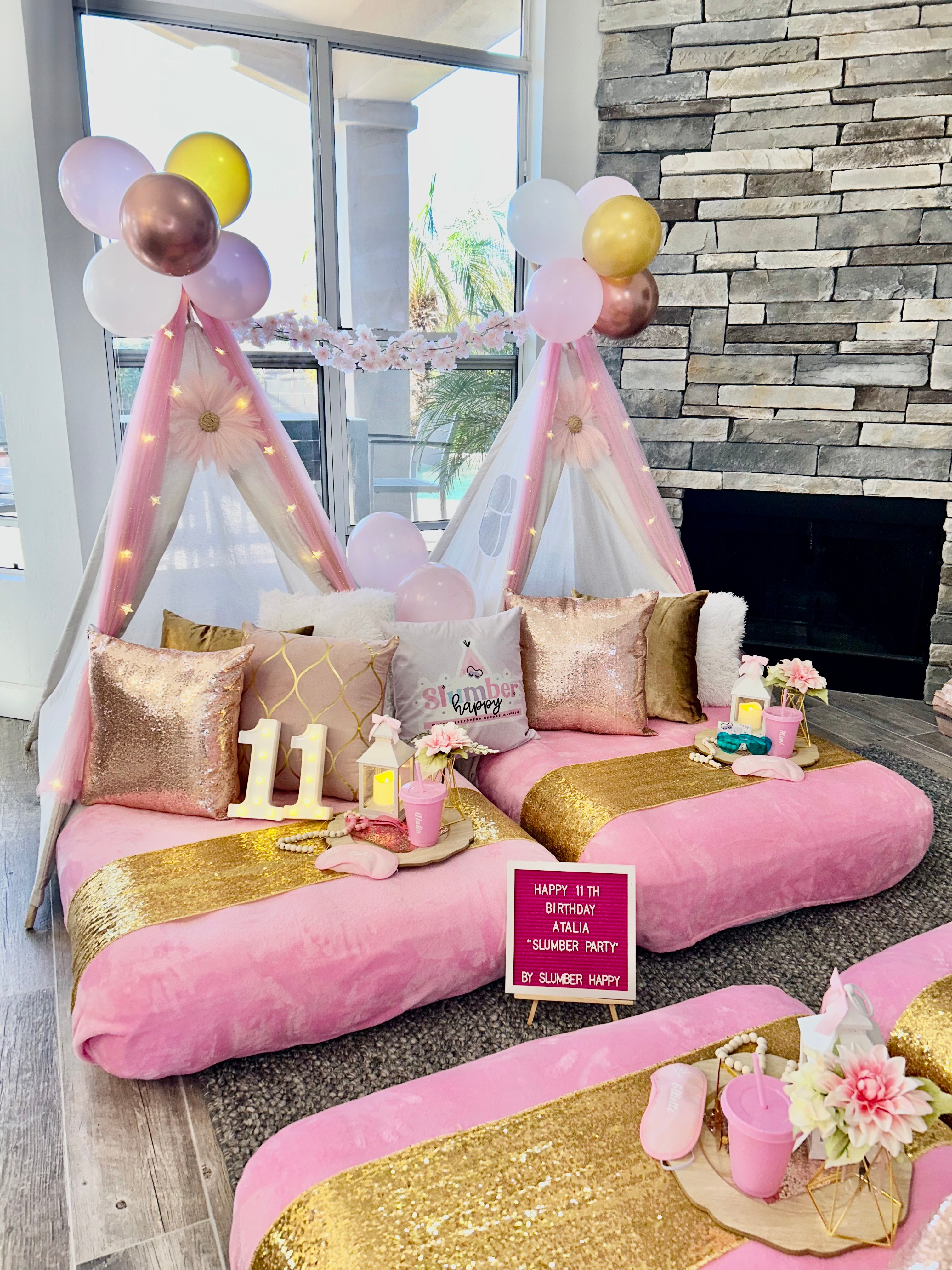 Decorative pink and gold setup with balloons, pillows, and a cake for a celebration, in a bright room with a stone wall.
