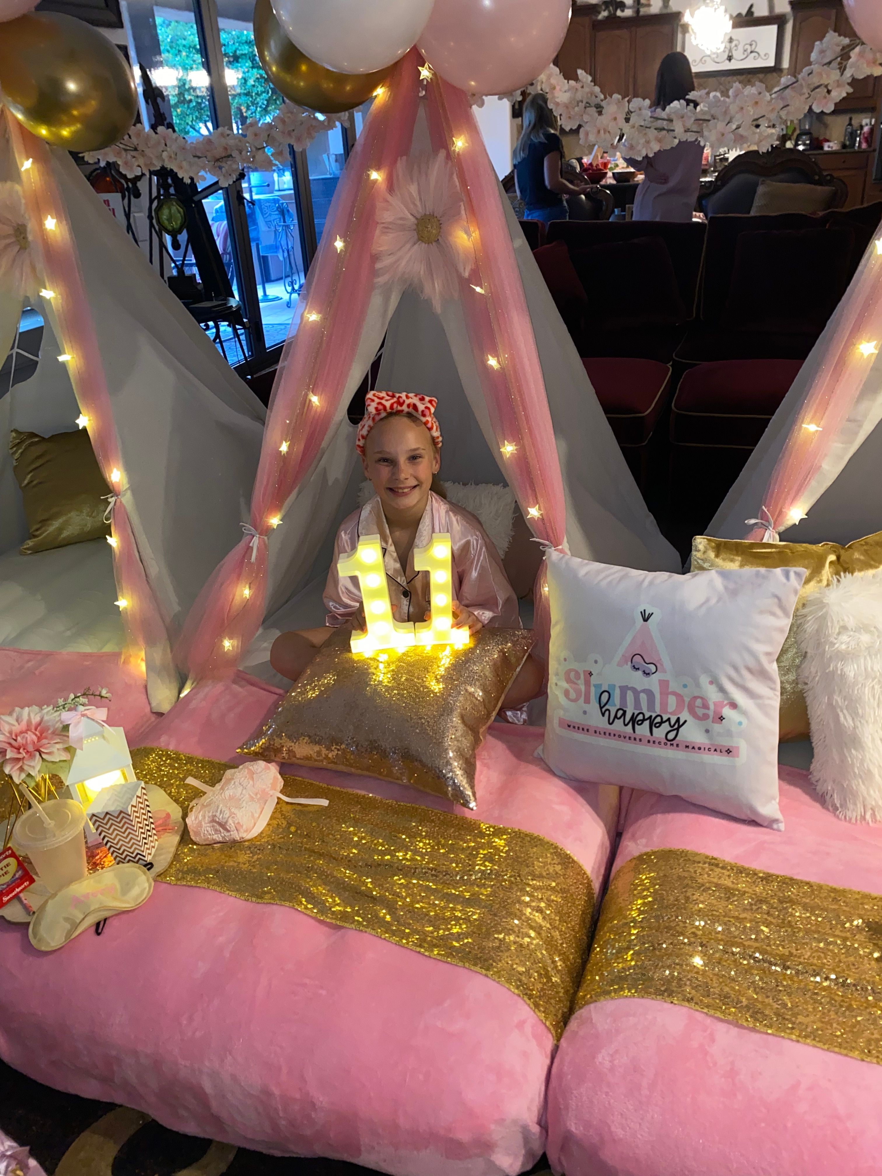 Girl sitting on decorated pink and gold bed with balloons and pillows, birthday celebration setup indoors.
