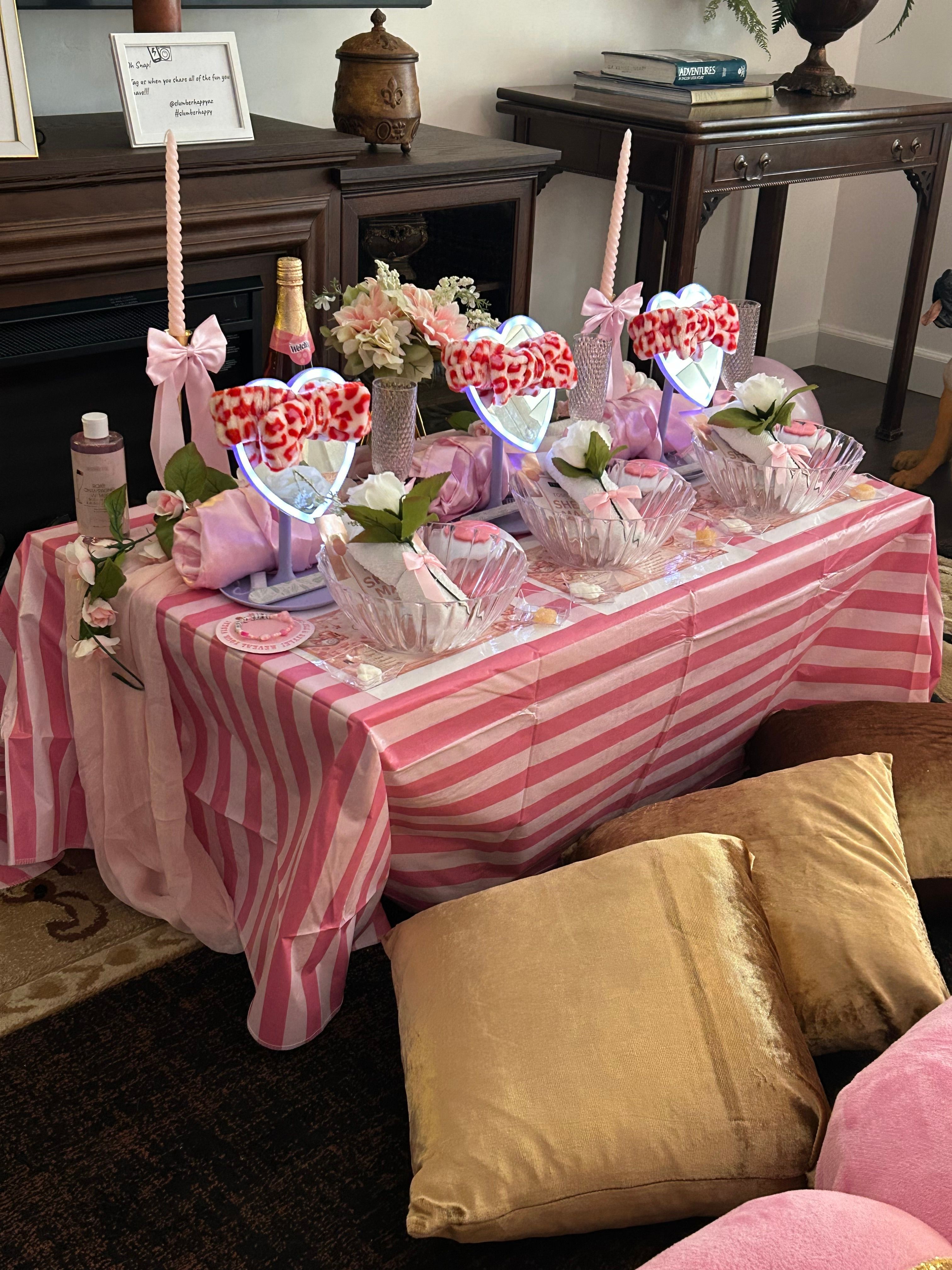Table with pink and white striped tablecloth, decorated with heart-shaped candies, flowers, and candles, in a room with furniture.