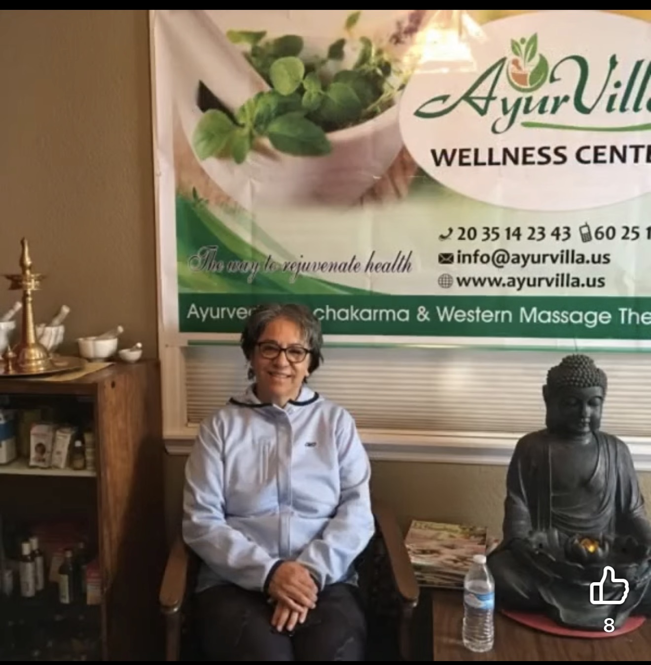 Woman sitting on chair next to a Buddha statue and a water bottle, in wellness center with banner behind.