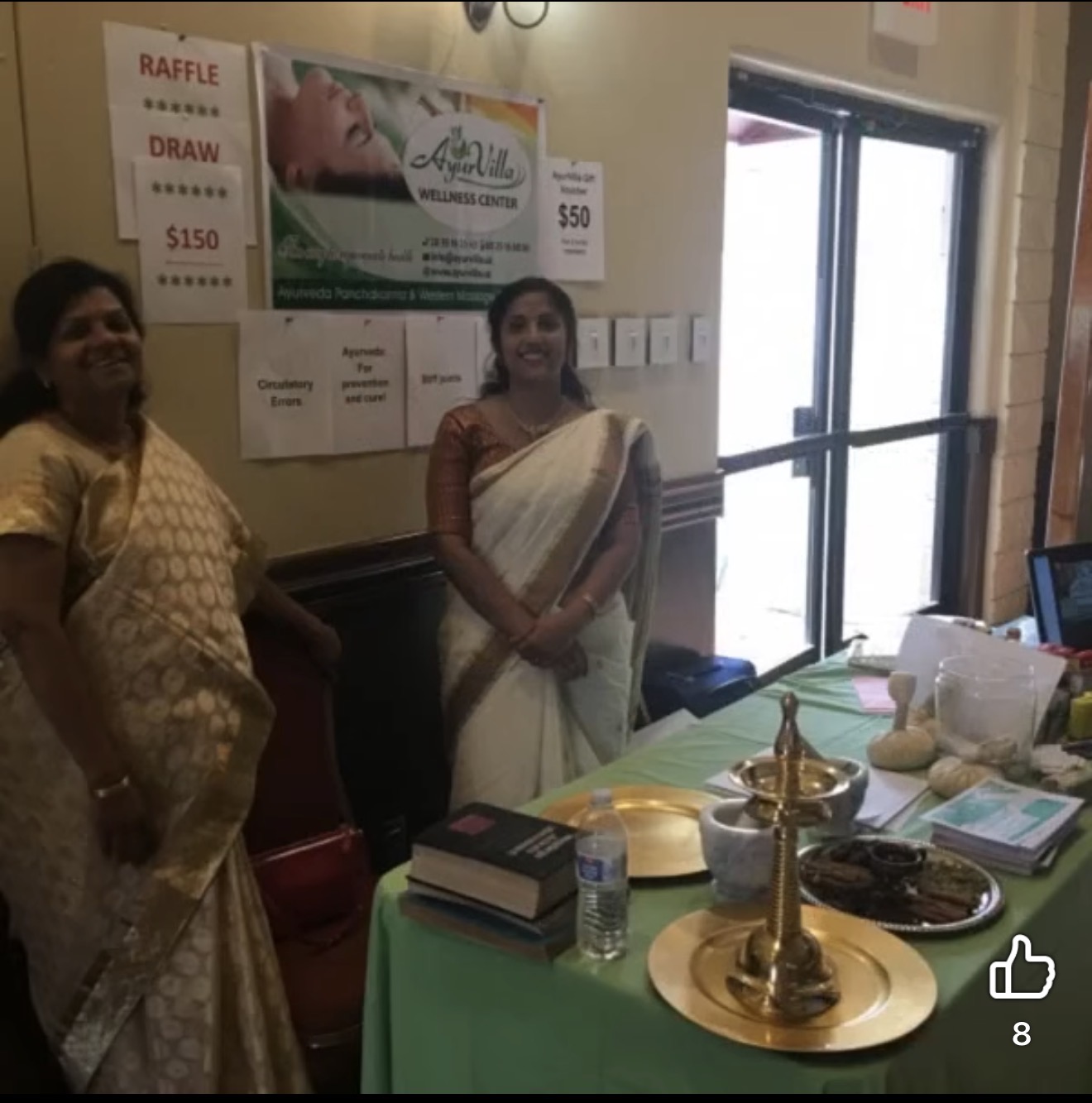 Two women in sarees standing near a table with books, a water bottle, and decorative items inside a room.