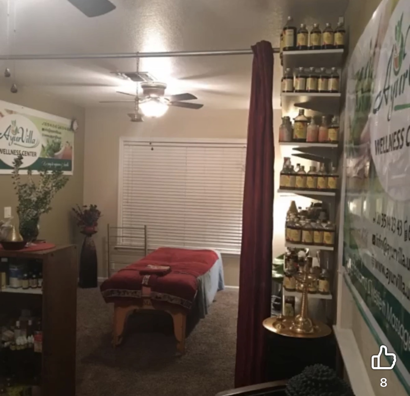 Room with massage table, shelves with jars, window with blinds, ceiling fan, and a curtain.