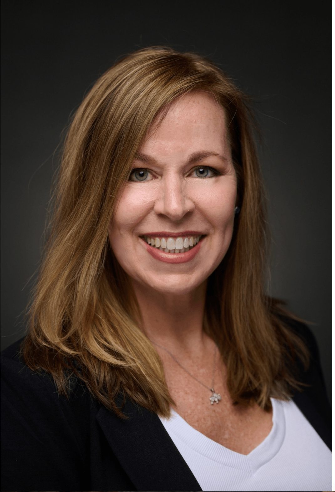 Woman with shoulder-length light brown hair smiling, wearing a black blazer and white top, against dark background.