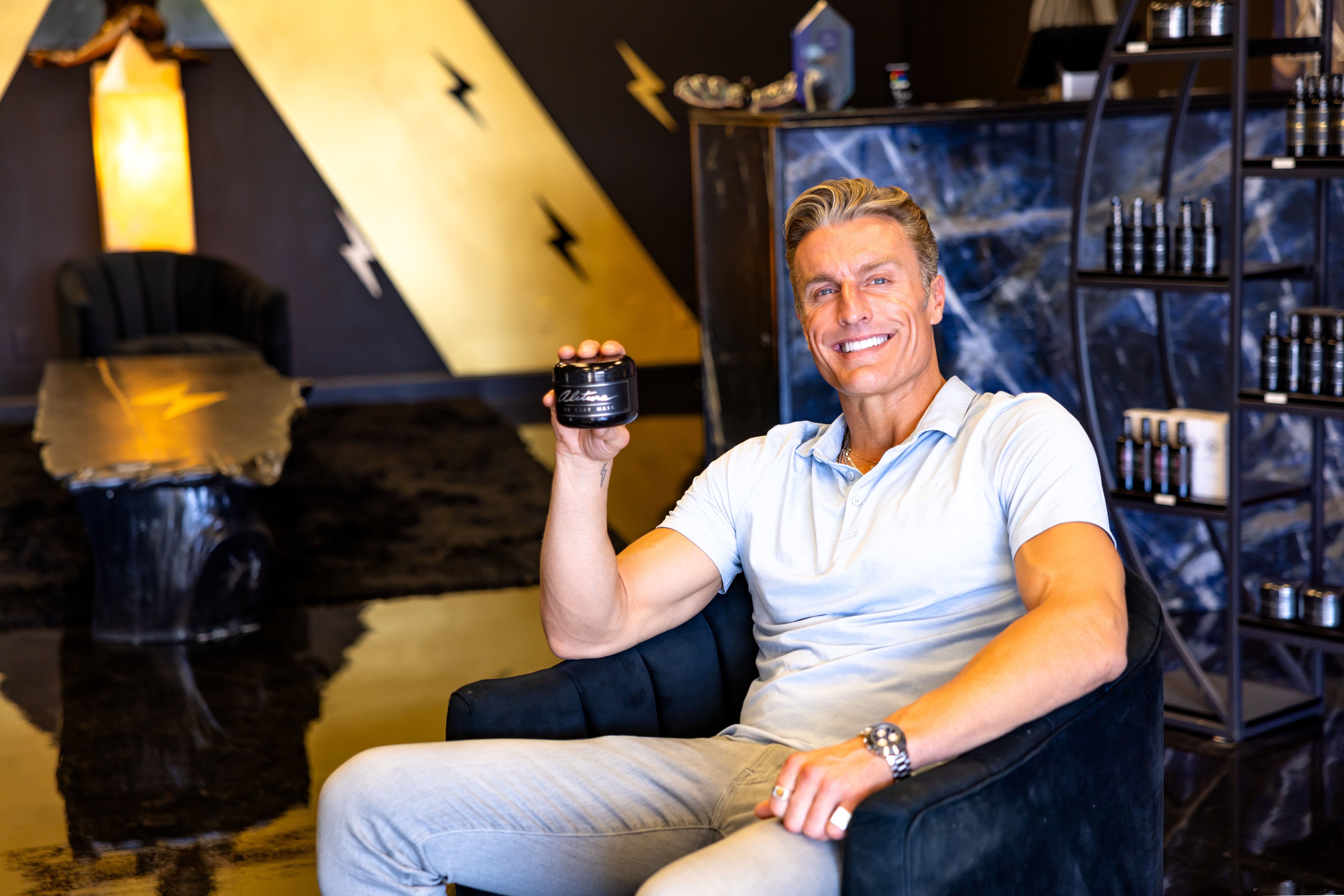 Smiling man sitting on a black chair holding a jar, in a room with dark walls and shelves.