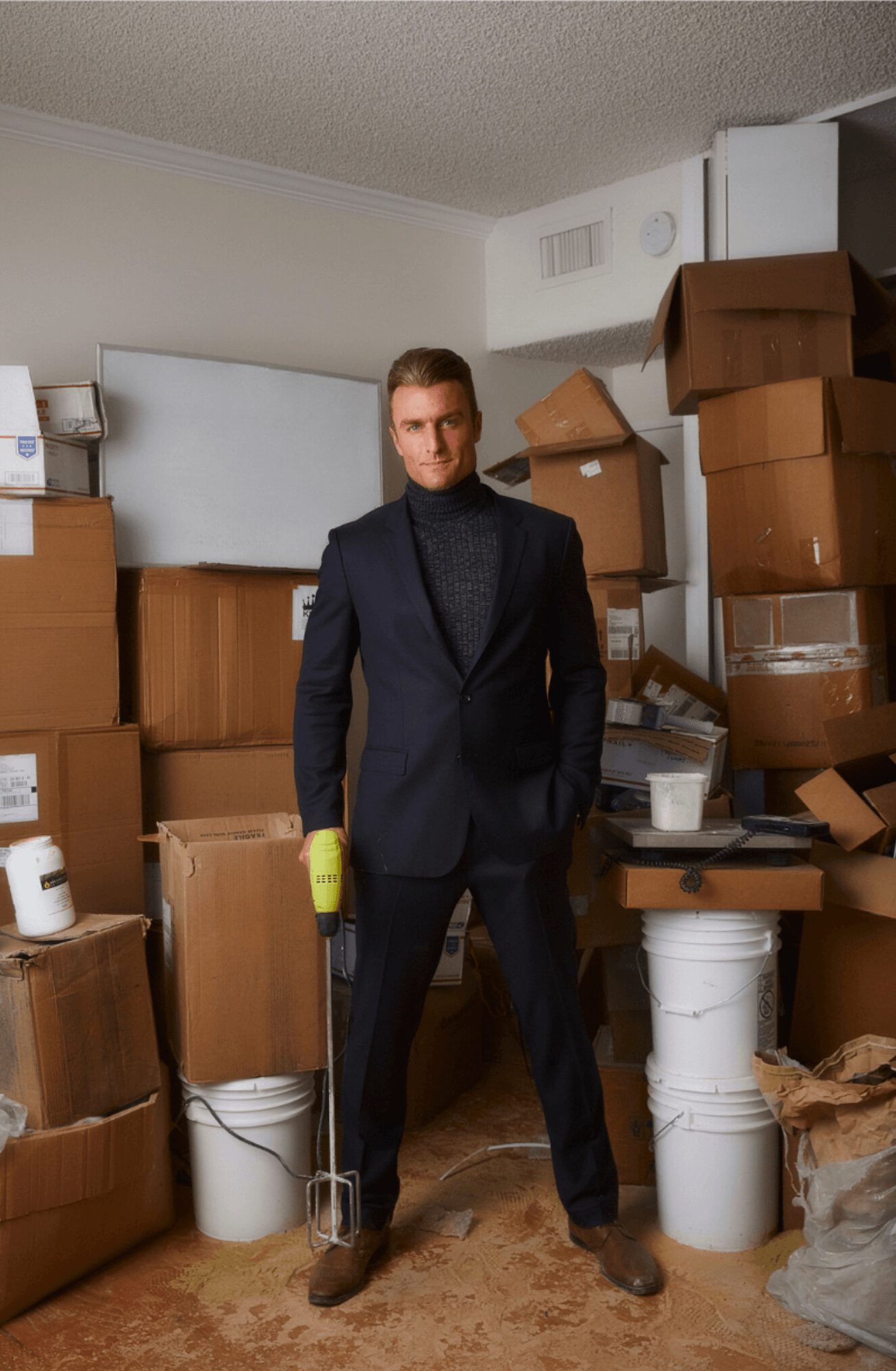 Man in a suit standing among stacked boxes and clutter in a room.