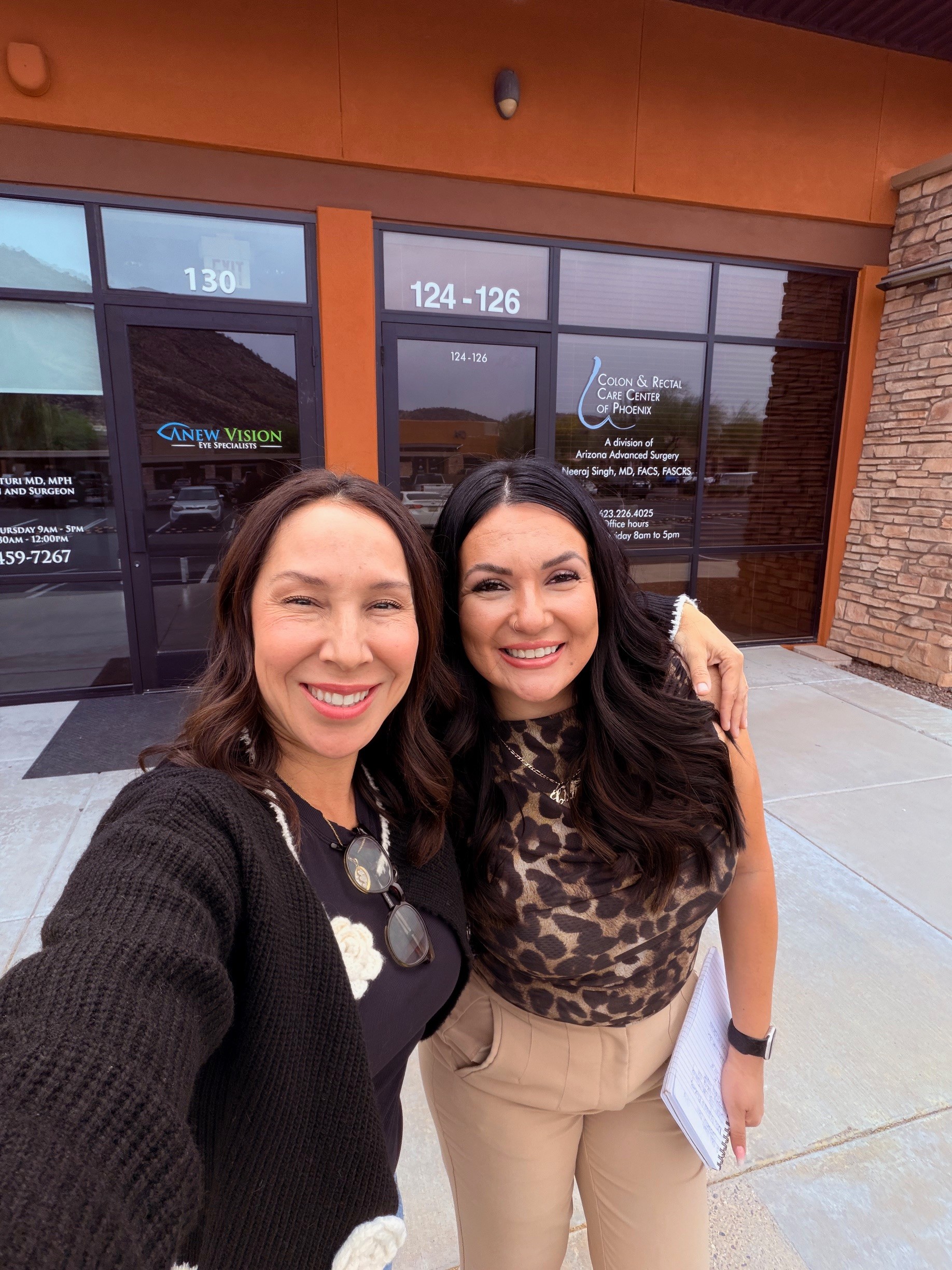 Two women smiling and taking a selfie outside a building with glass doors and brick wall.
