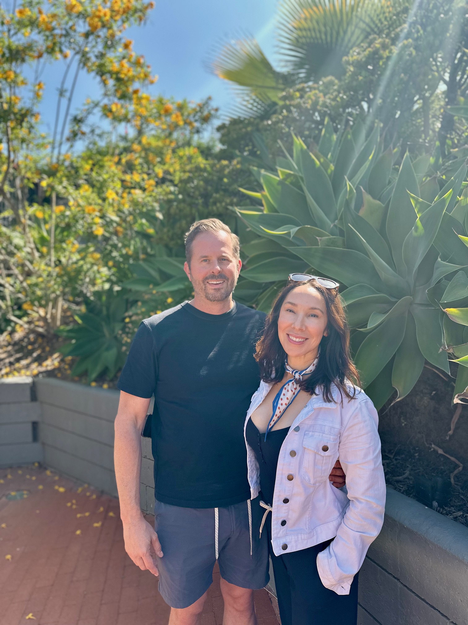 A man and woman stand outdoors near large green plants, smiling at the camera on a sunny day.