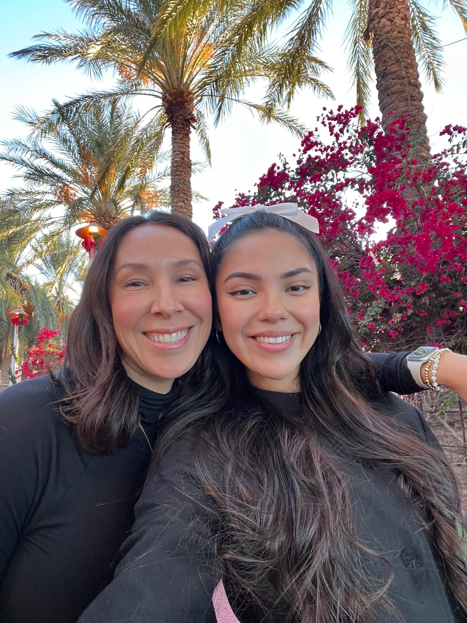Two women smiling outdoors with palm trees and pink flowering bushes in background.