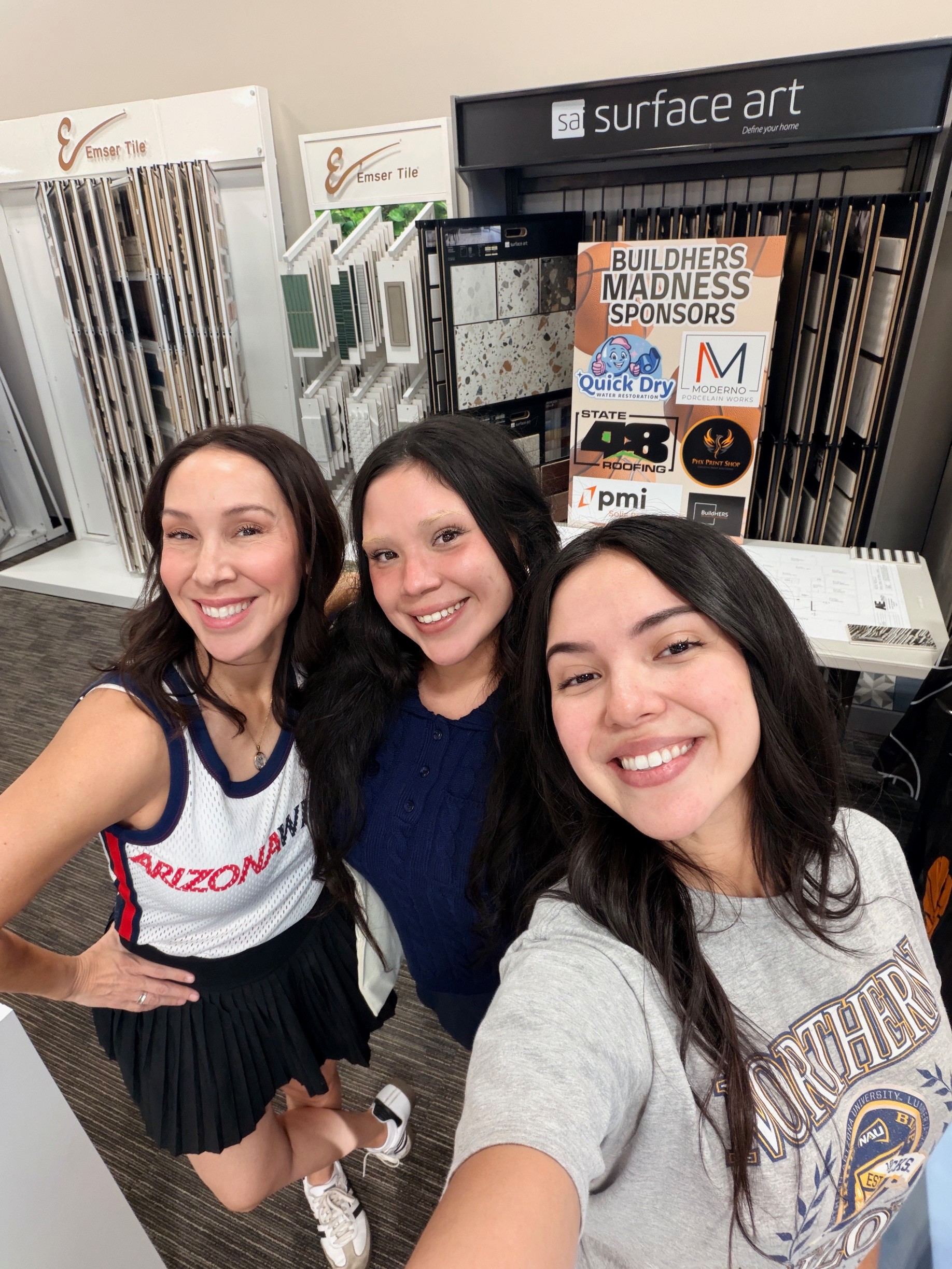 Three smiling women taking a selfie inside a store with building materials and signage in the background.