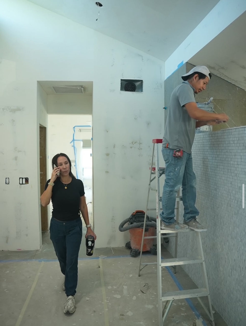Woman walking with a drink in hand, man on ladder working on wall, interior space under renovation.
