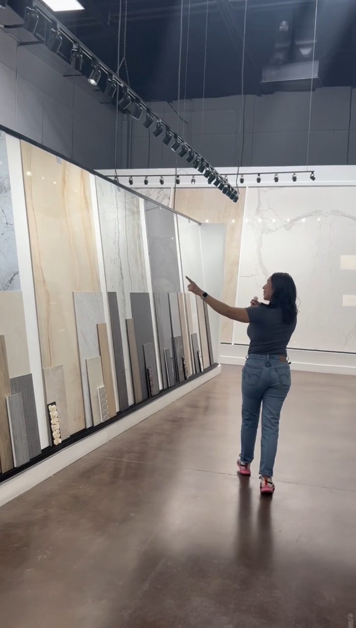 Woman in jeans and black top points at large display of various tile samples in a showroom.