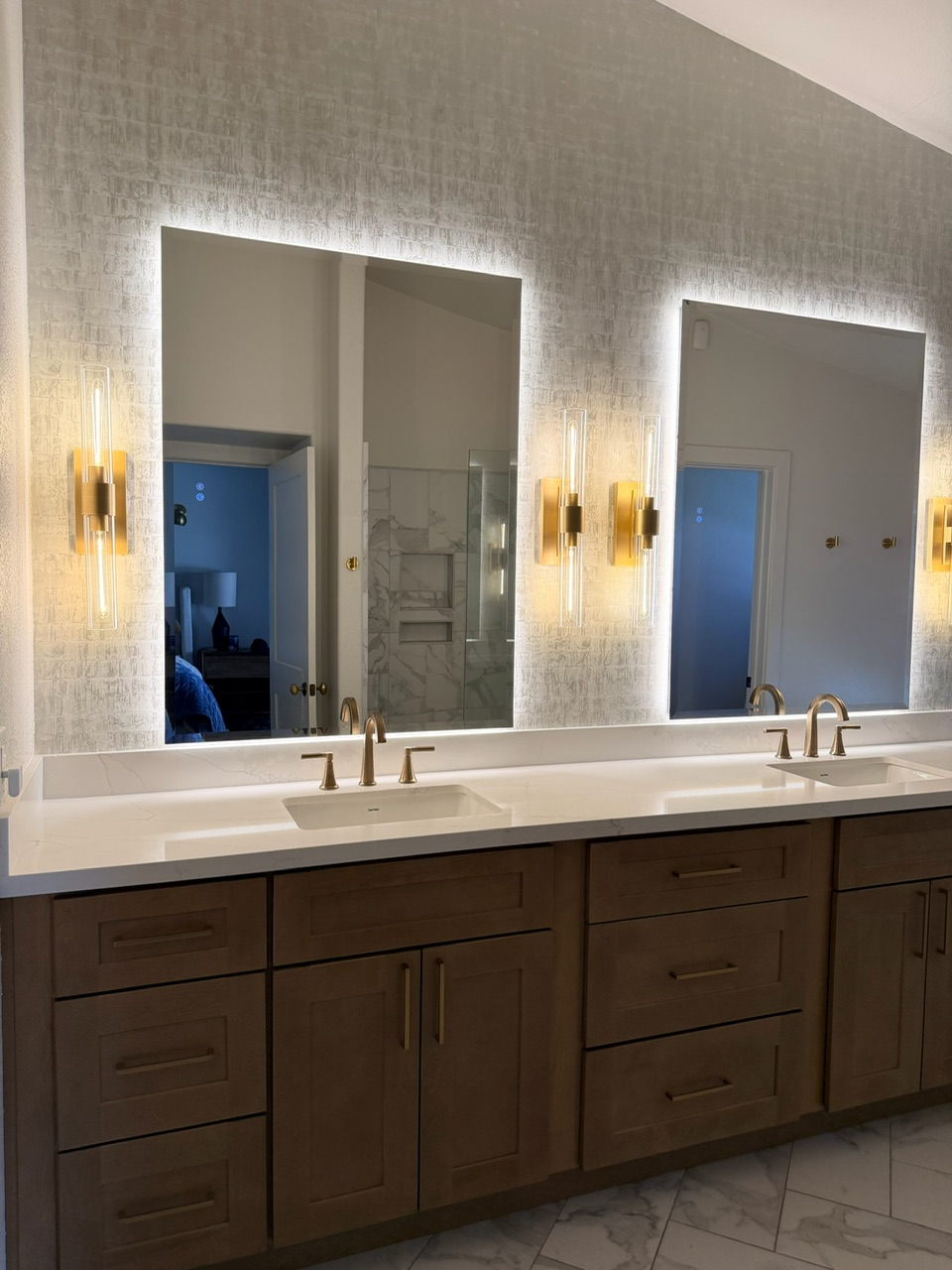 Double bathroom mirrors with backlit lighting, gold fixtures, wooden cabinet, and tiled wall and floor.