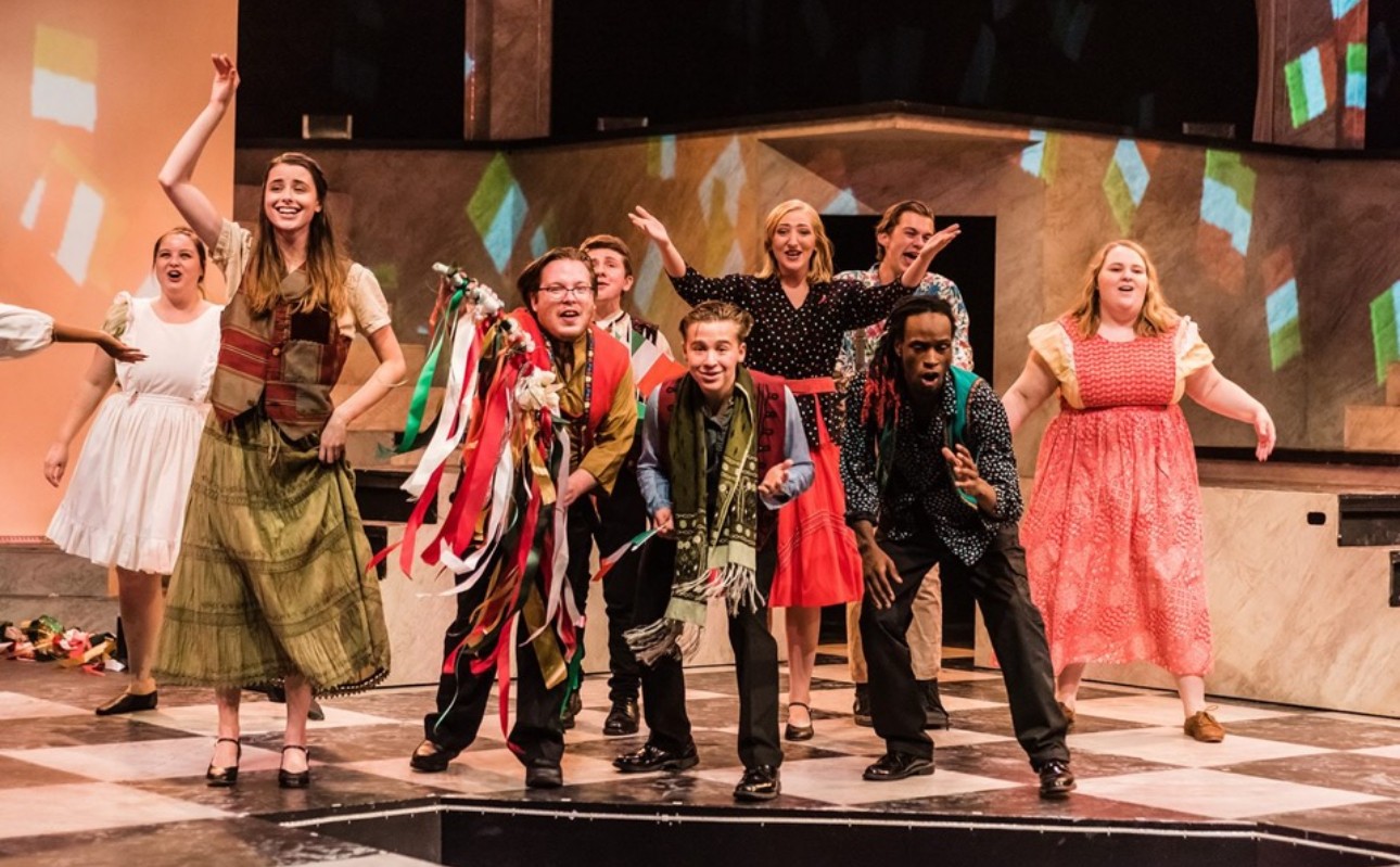 Group of children and adults dancing on stage with colorful background and checkered floor.