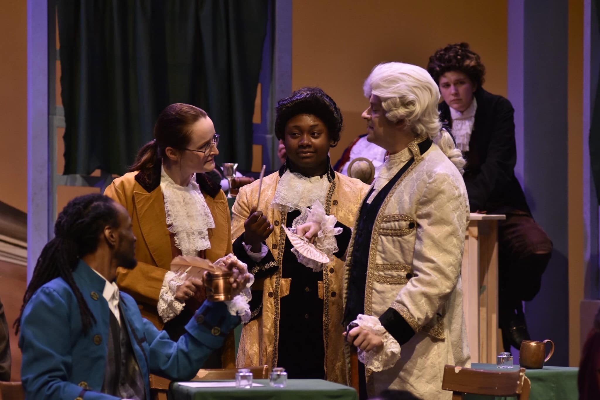 Four people in period costumes engaged in conversation on a stage with a woman in the background.