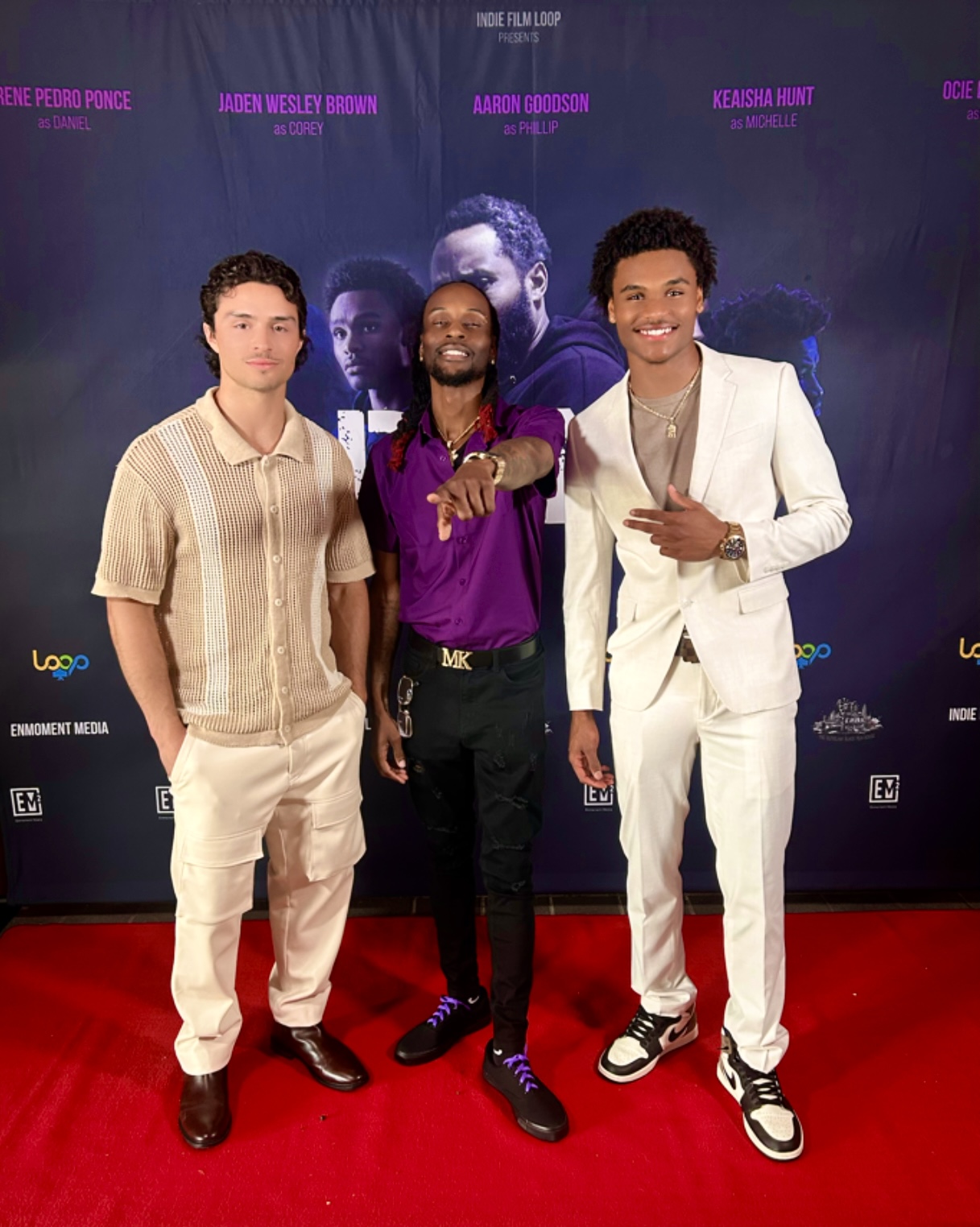 Three men standing on red carpet in front of a dark backdrop with images and text, smiling at camera.