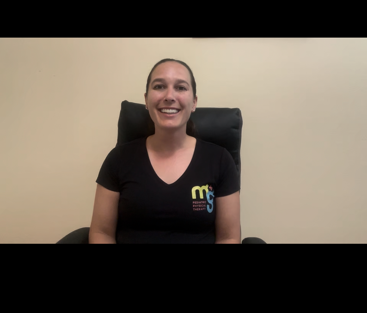 Smiling woman sitting in a chair against a plain wall, wearing a black shirt with colorful logo, facing forward.