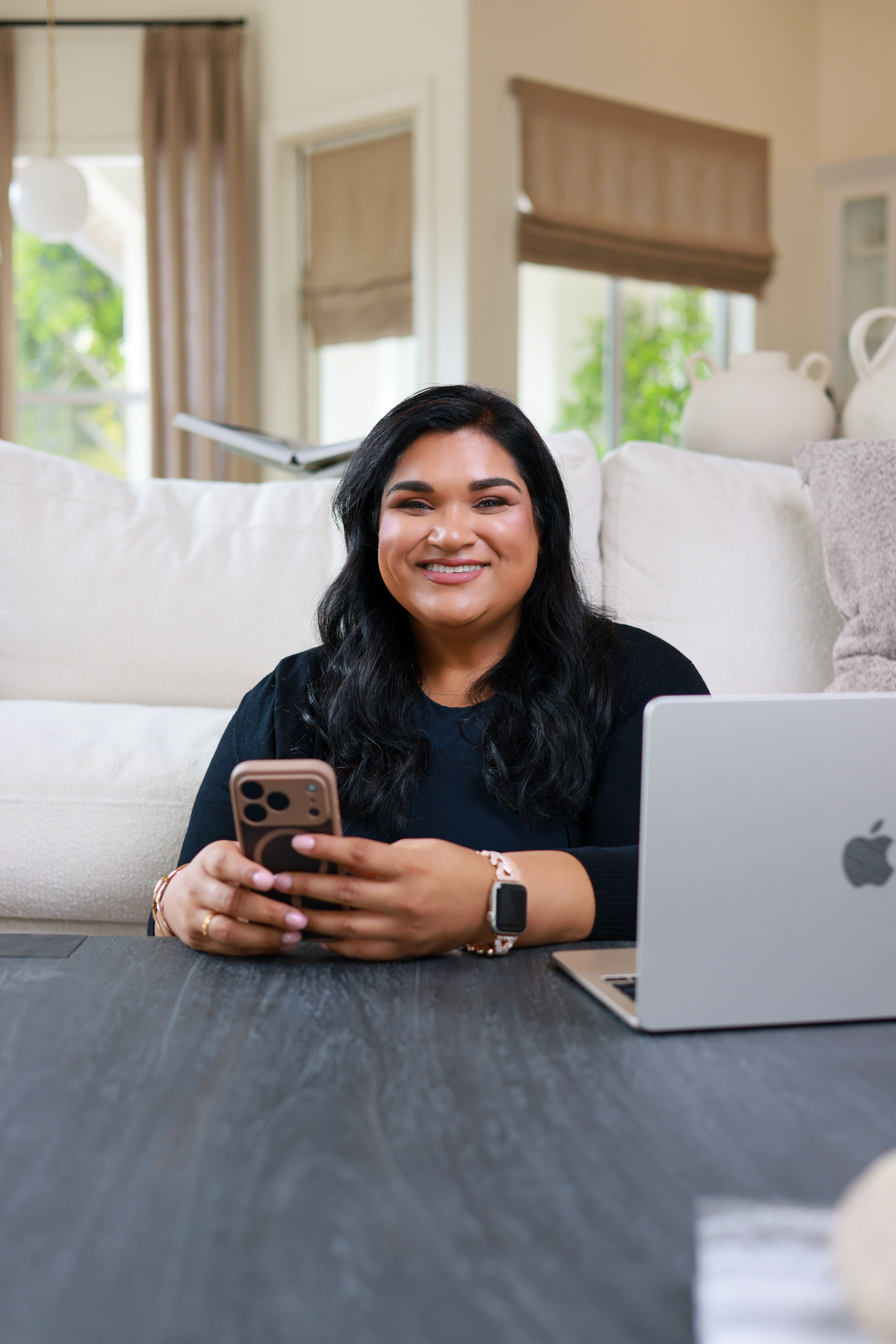 Woman with dark hair smiling, holding a smartphone, sitting at a table with a laptop, in a bright living room.