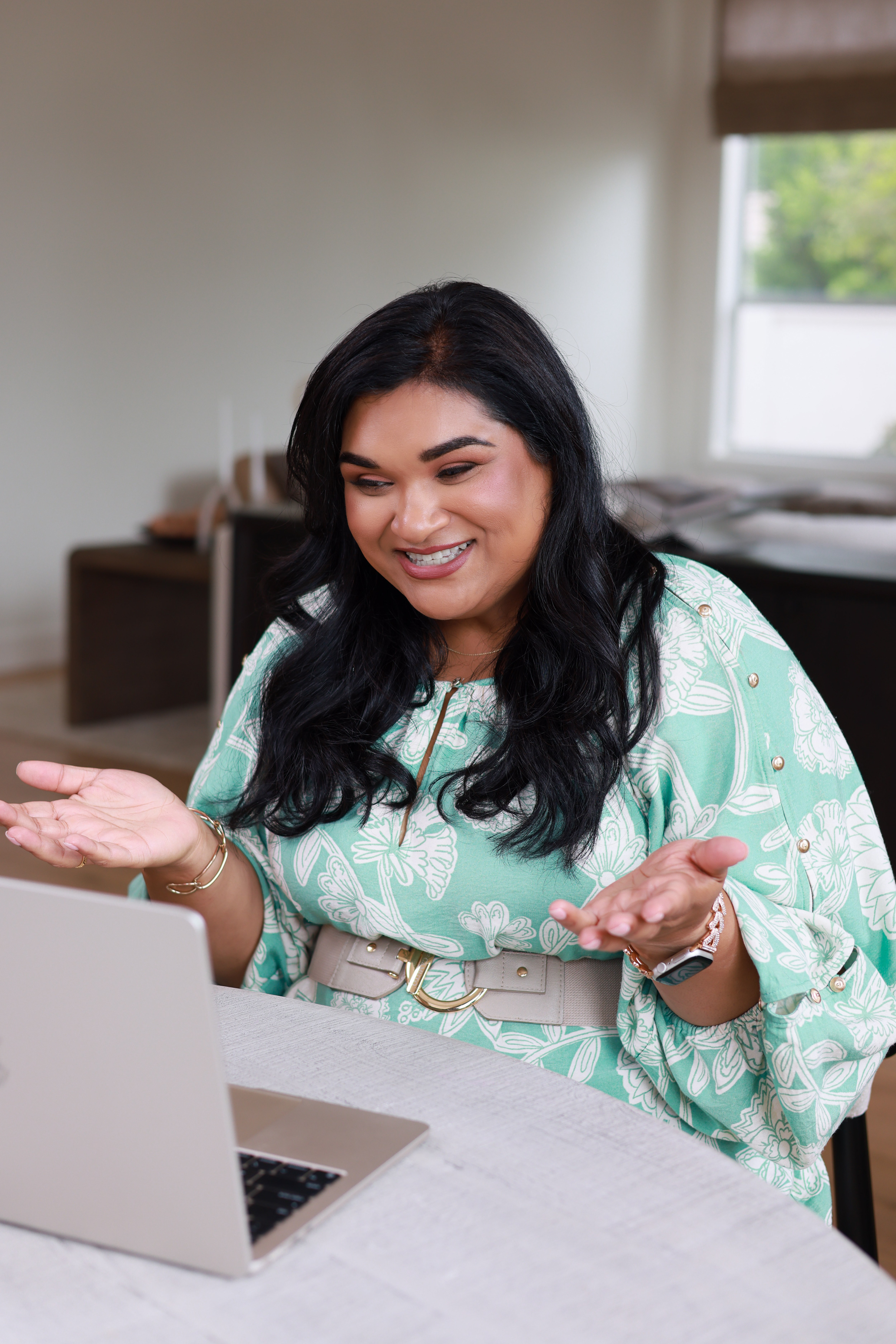 Woman with dark hair smiling and gesturing while looking at a laptop, sitting indoors near a window.