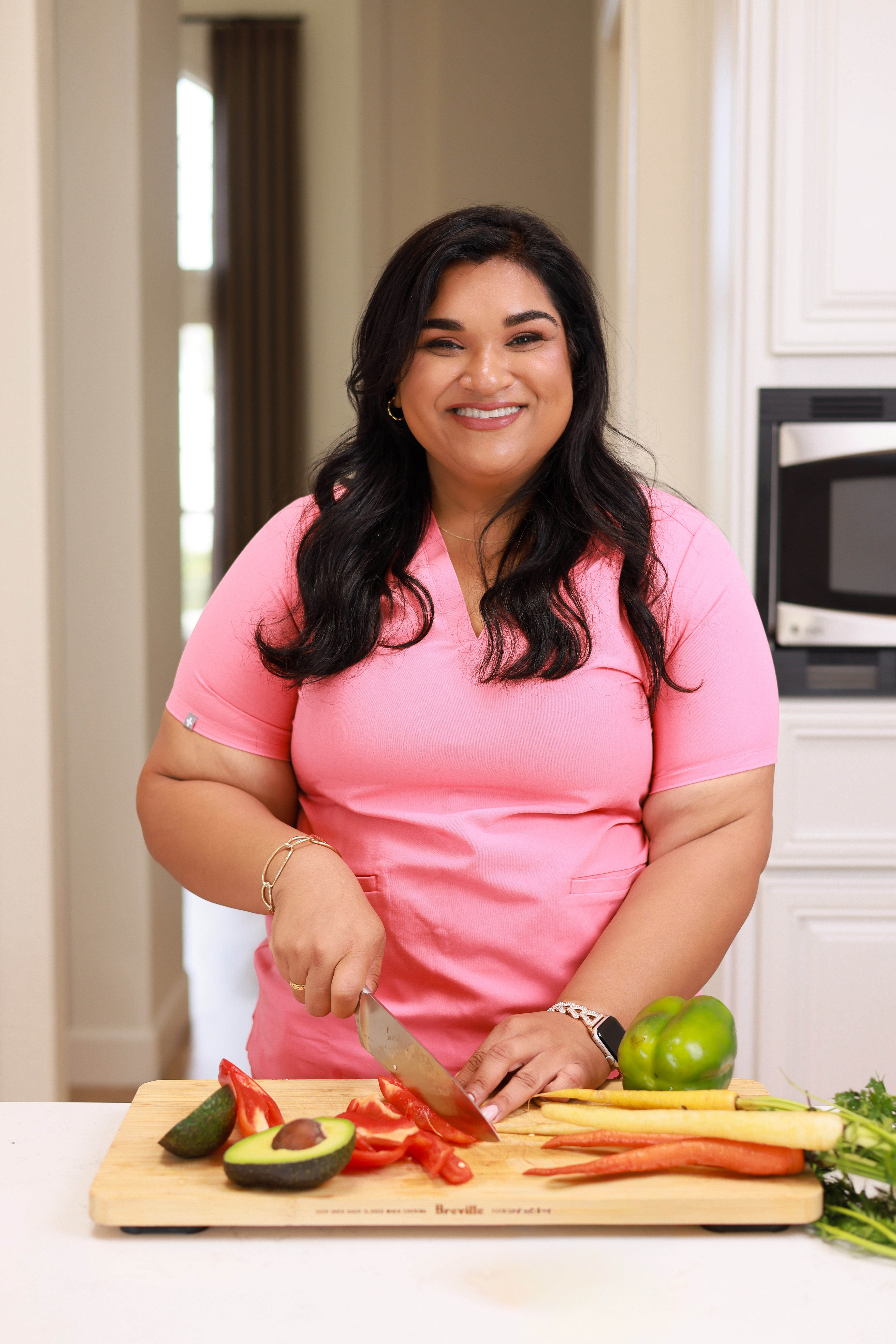 Woman with long dark hair in pink shirt chopping vegetables in a kitchen, smiling, with a microwave and white cabinets behind.