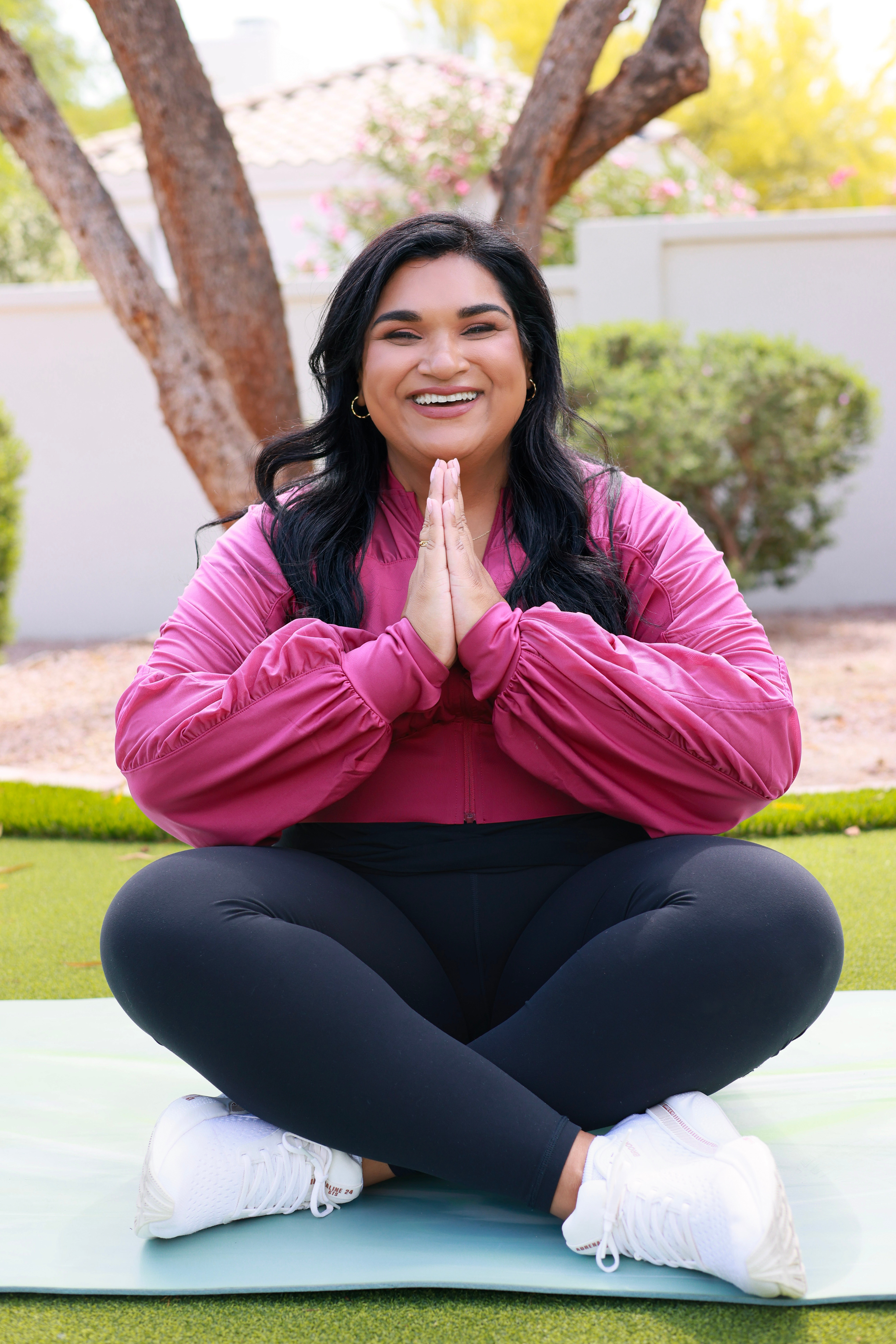 Woman sitting cross-legged outdoors, smiling, hands pressed together in a prayer gesture, trees and greenery in background.