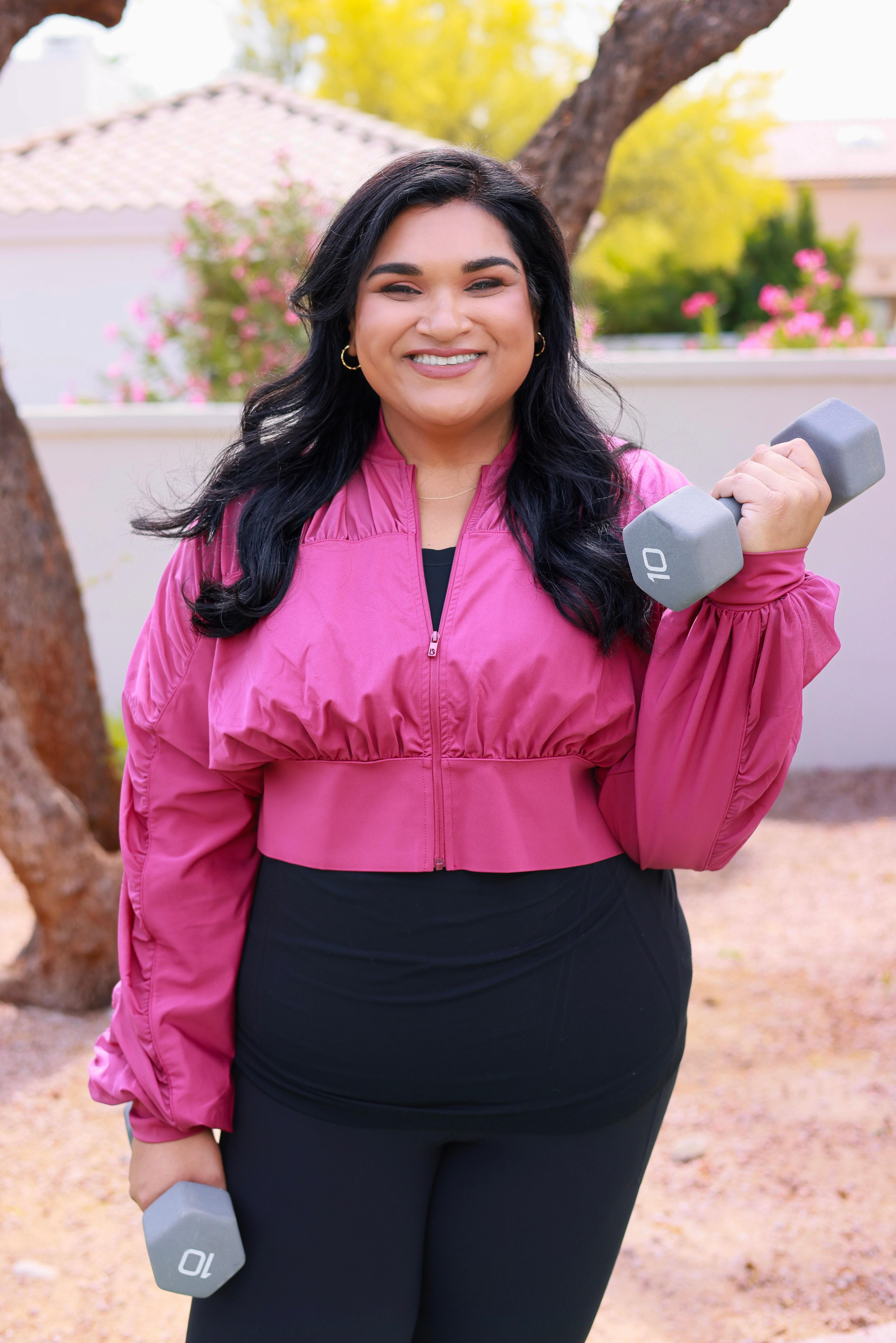 Woman smiling outdoors holding a gray dumbbell in her right hand, wearing a pink jacket and black pants.