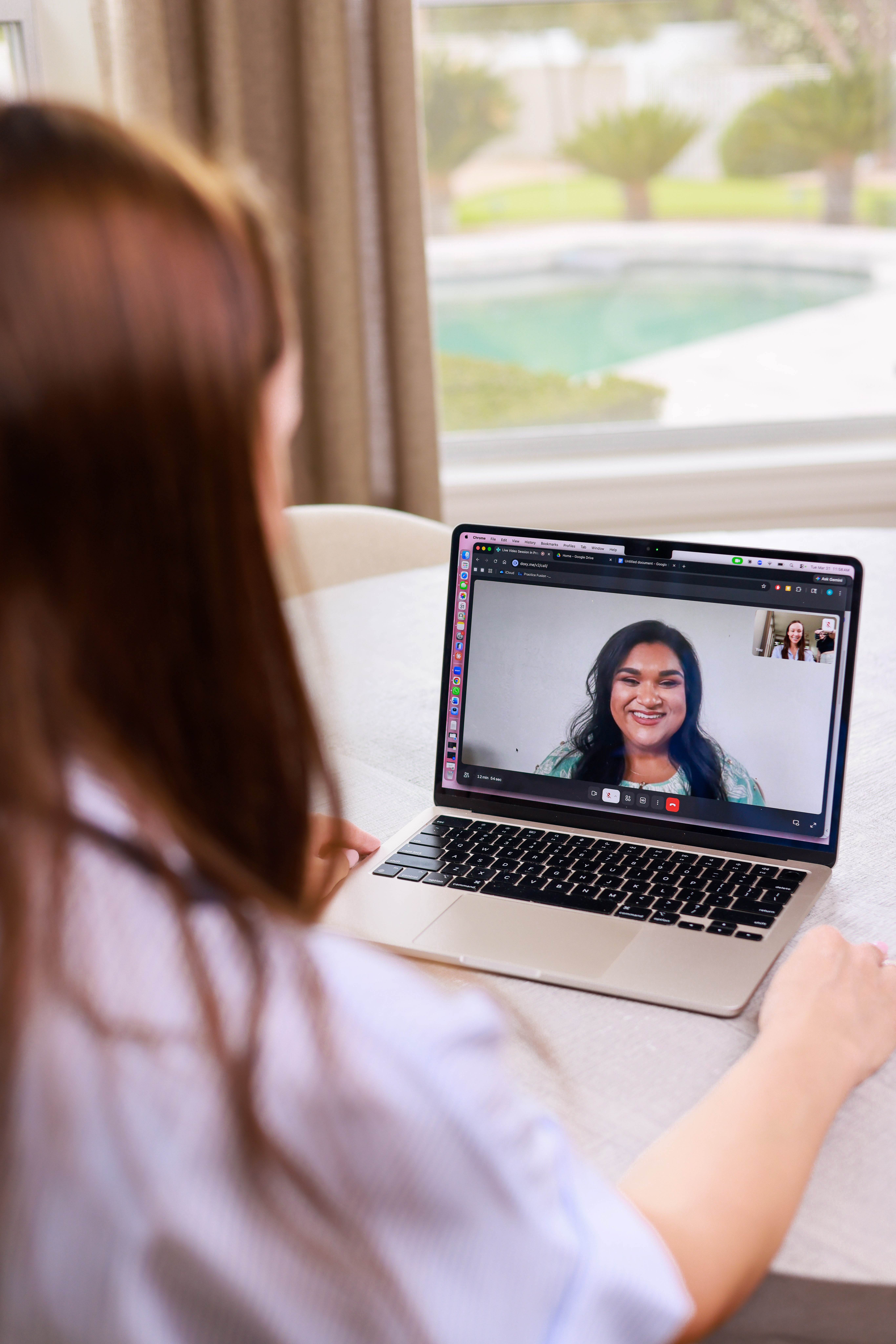 Person with long hair participating in a video call on a laptop, with a pool and garden visible outside window.