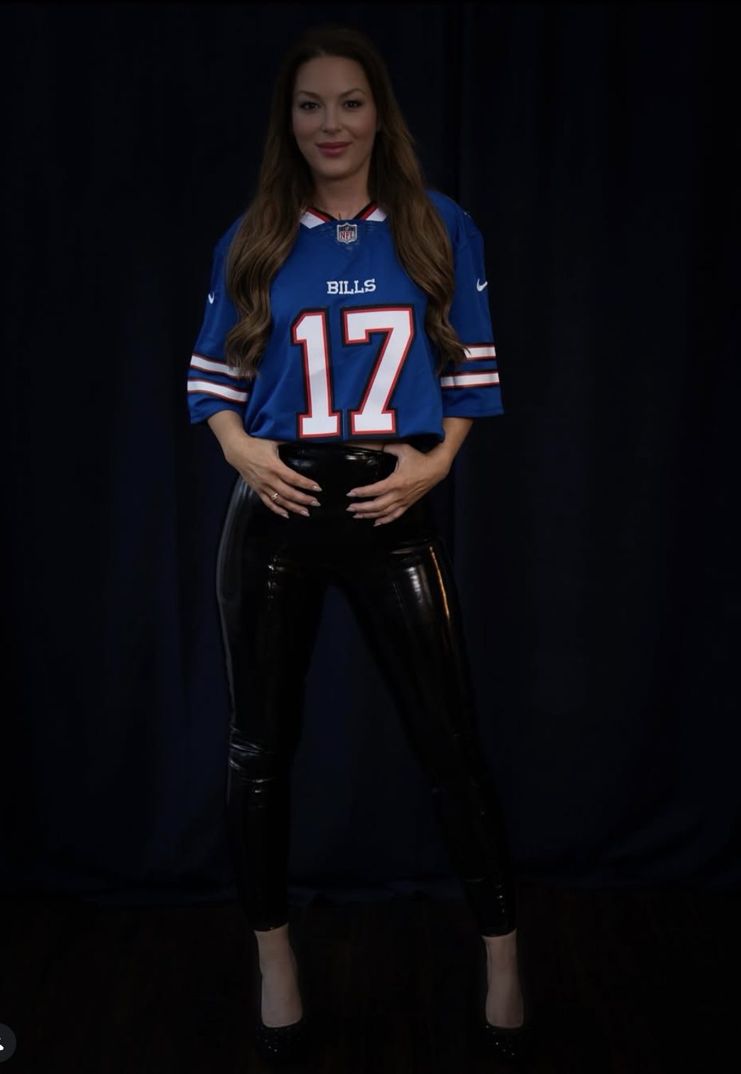 Young woman in a blue sports jersey with the number 17, black shiny pants, standing against a dark background.