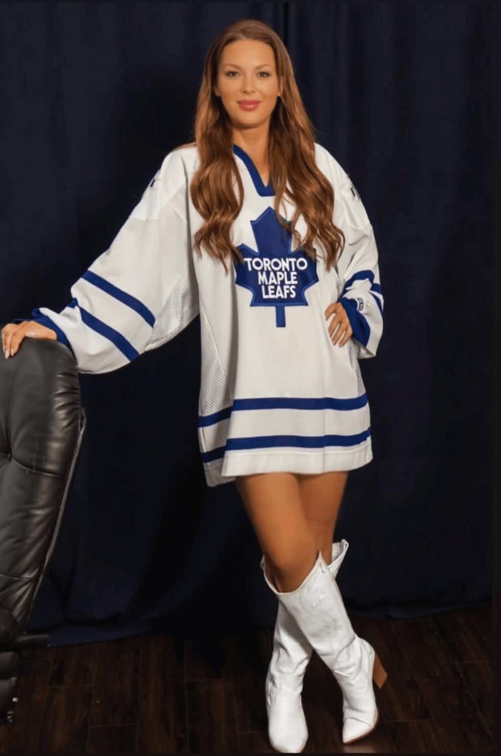 Woman in Toronto Maple Leafs hockey jersey and white boots standing indoors, dark curtain background.