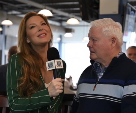 A woman with long red hair holding a microphone and smiling, talking to an older man with white hair in a casual setting.