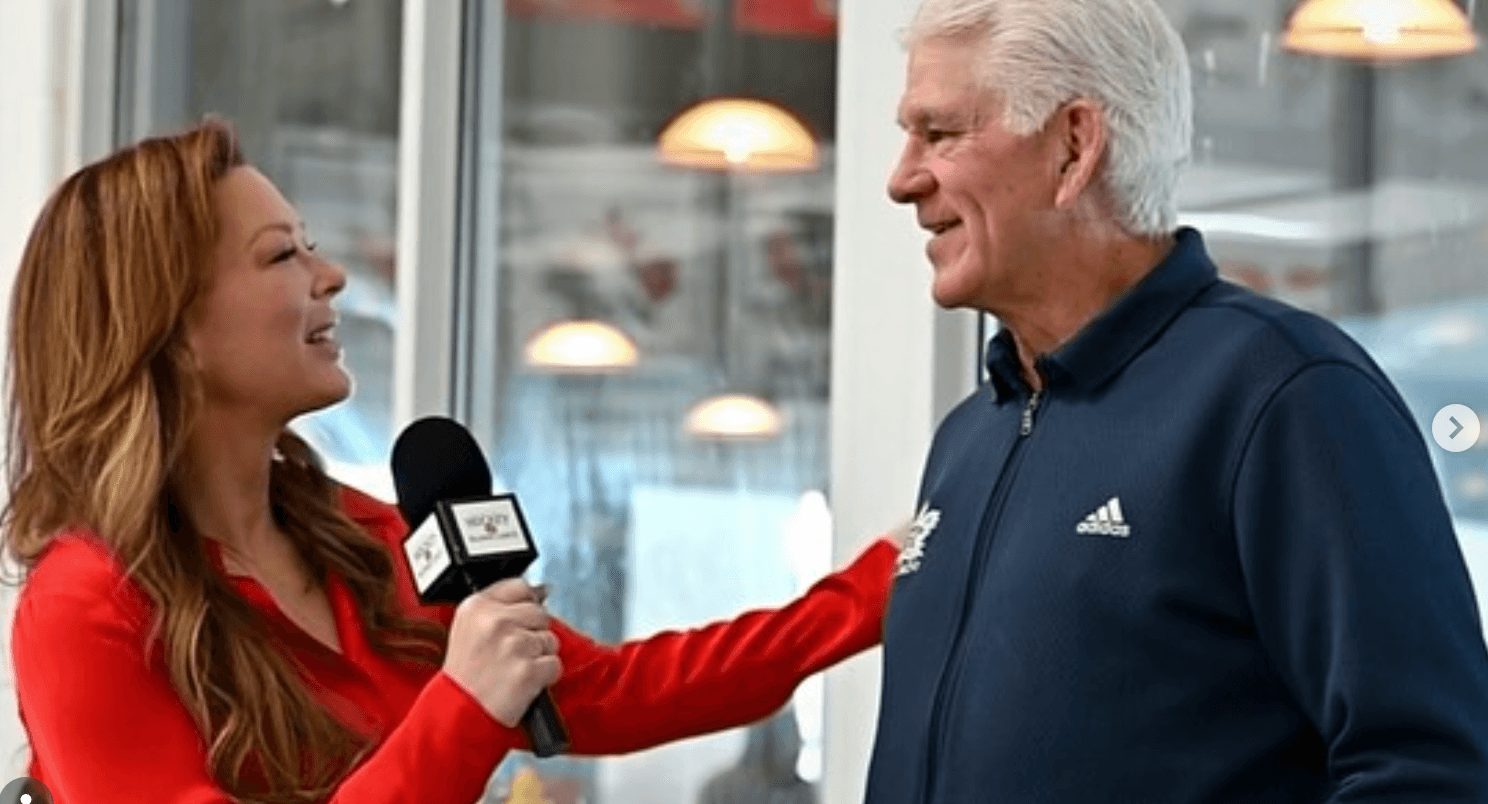 A woman with long hair holding a microphone interviews an older man with white hair, both smiling, indoors.