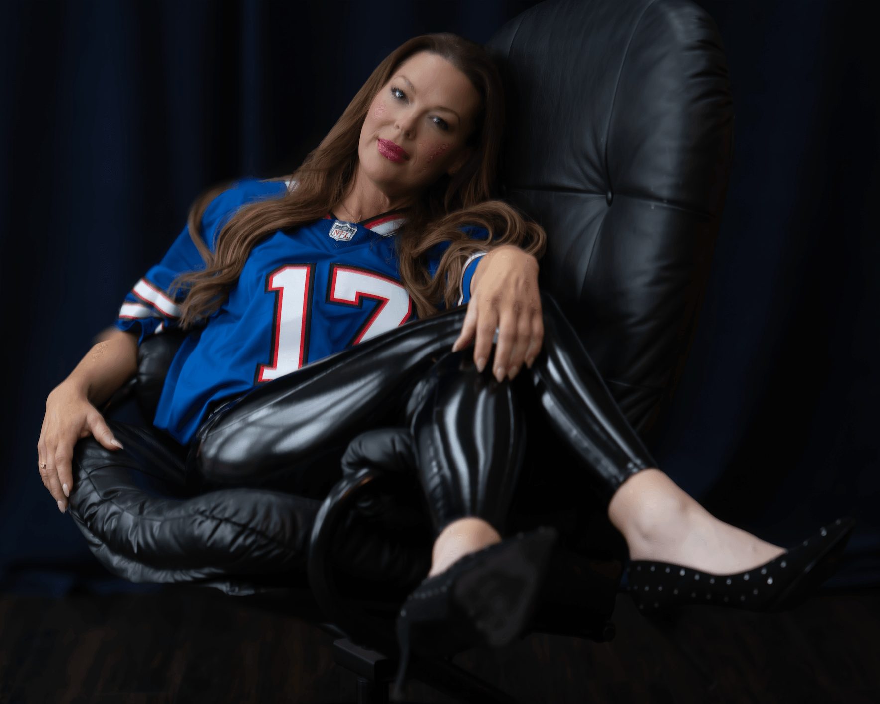 Woman with long hair wearing a blue sports jersey and black shiny pants, sitting on a black chair with dark curtains behind her.