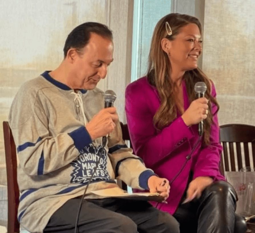 Two people sitting and speaking into microphones, one wearing a hockey jersey, the other in a pink blazer, smiling.
