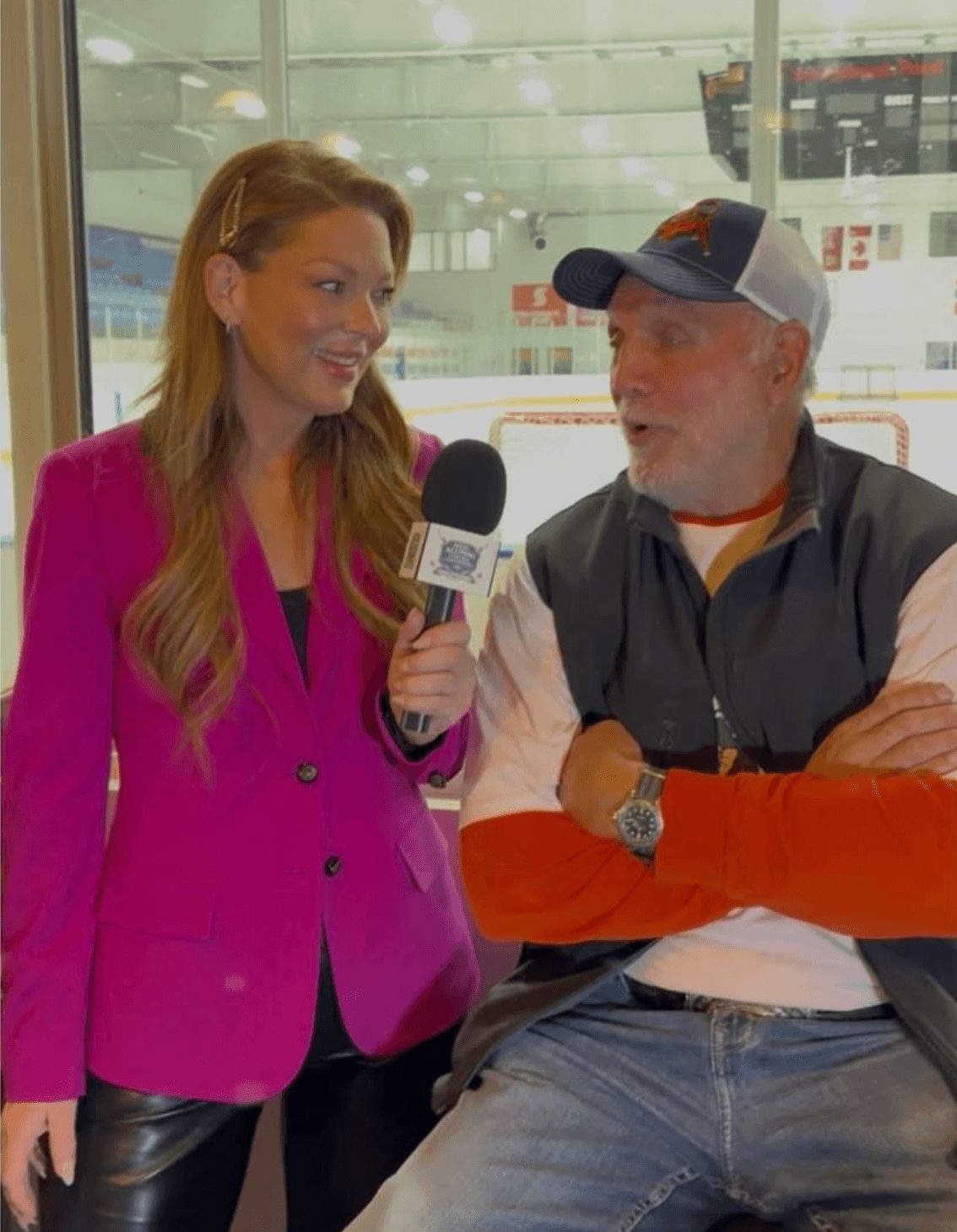 Woman in pink blazer holding microphone interviews man in cap at ice rink, smiling, background shows ice and rink boards.