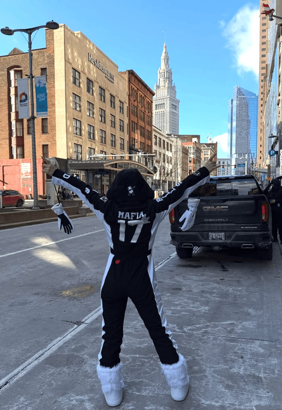 Person standing on city street with arms raised, wearing black hoodie and white shoes, tall building in background.