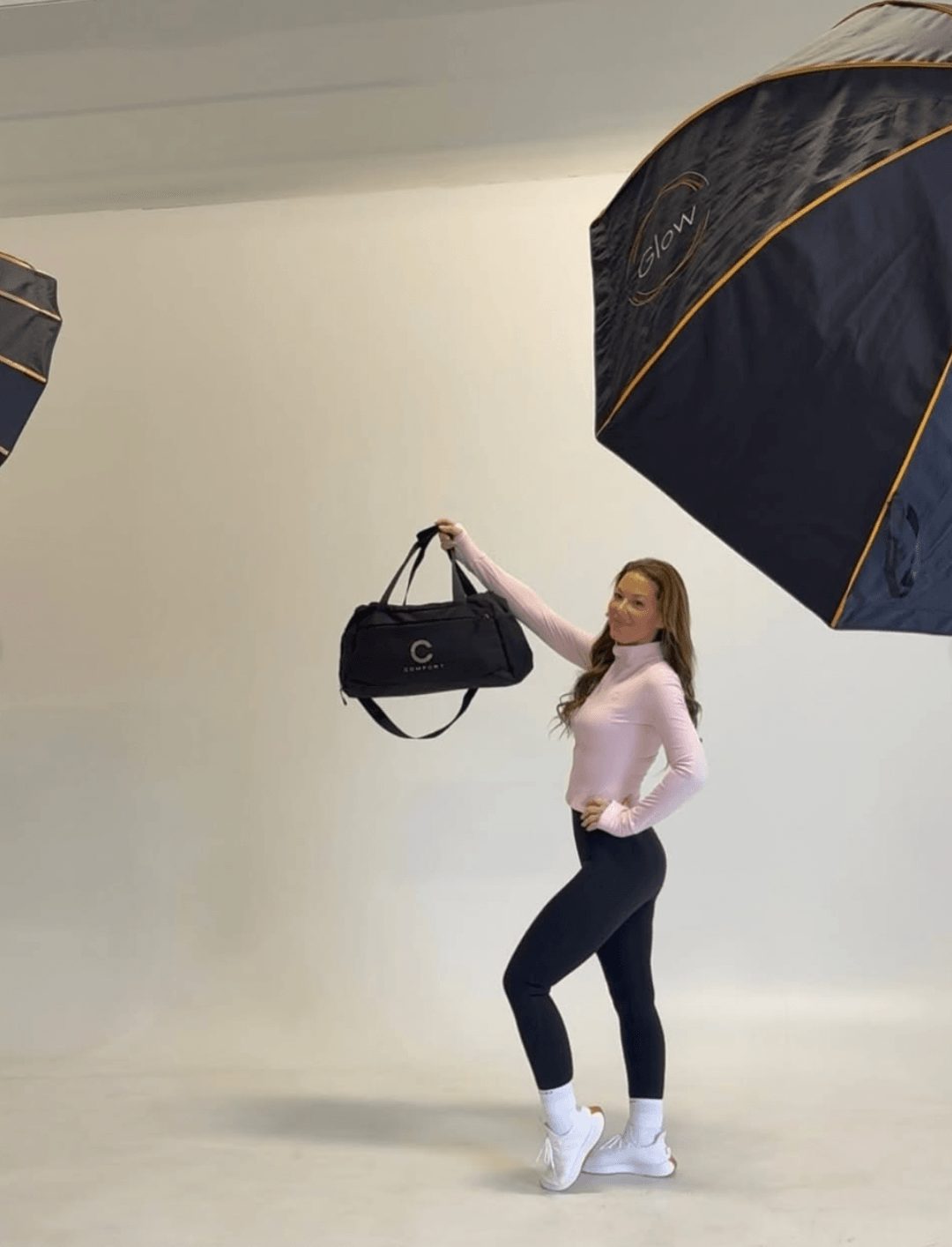 Woman standing in a studio with large photography umbrellas, holding a black bag, wearing a pink top, dark pants, and white sneakers.