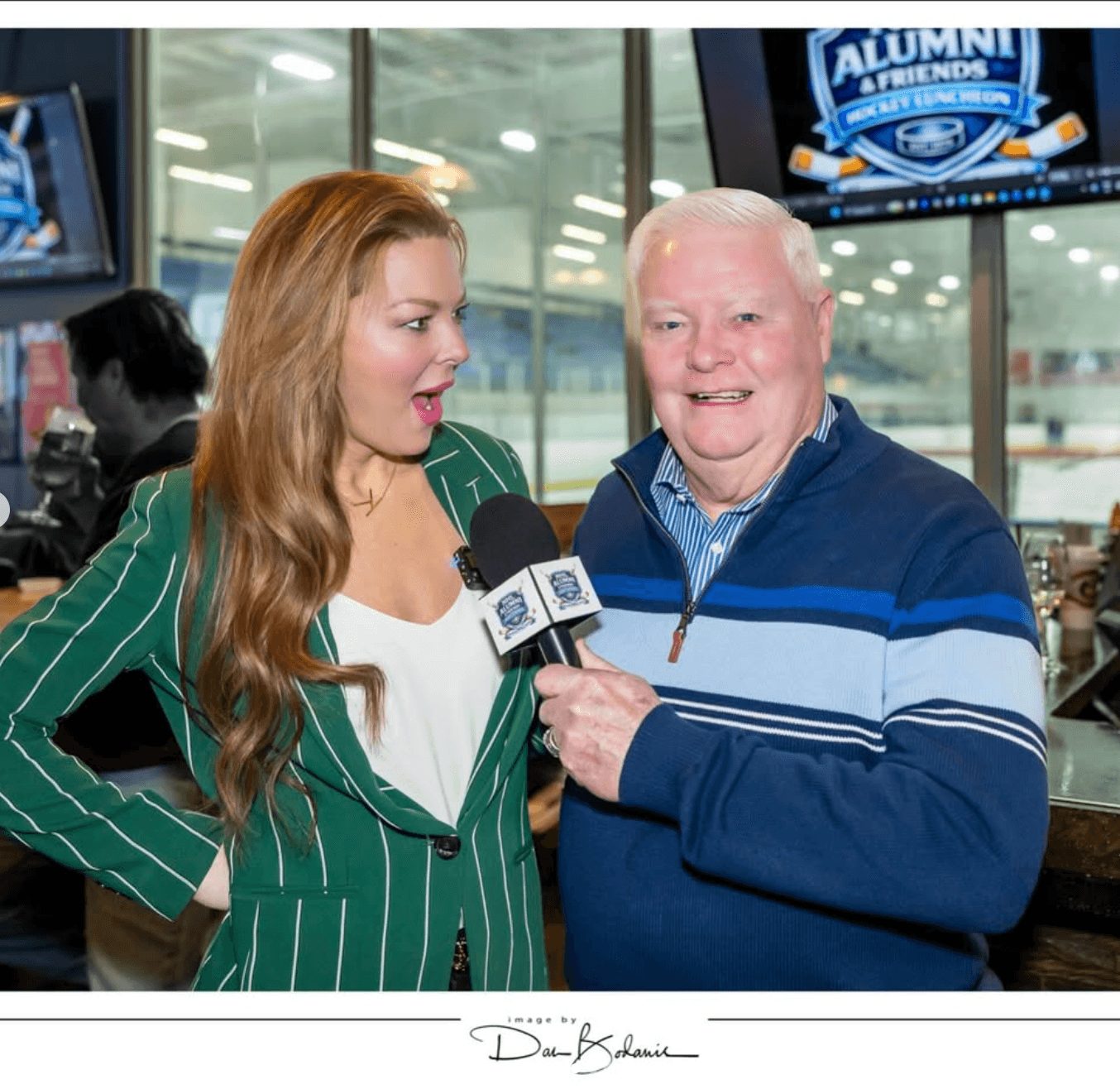 Woman with long hair and green striped blazer speaks into microphone held by smiling man in blue sweater, indoors.