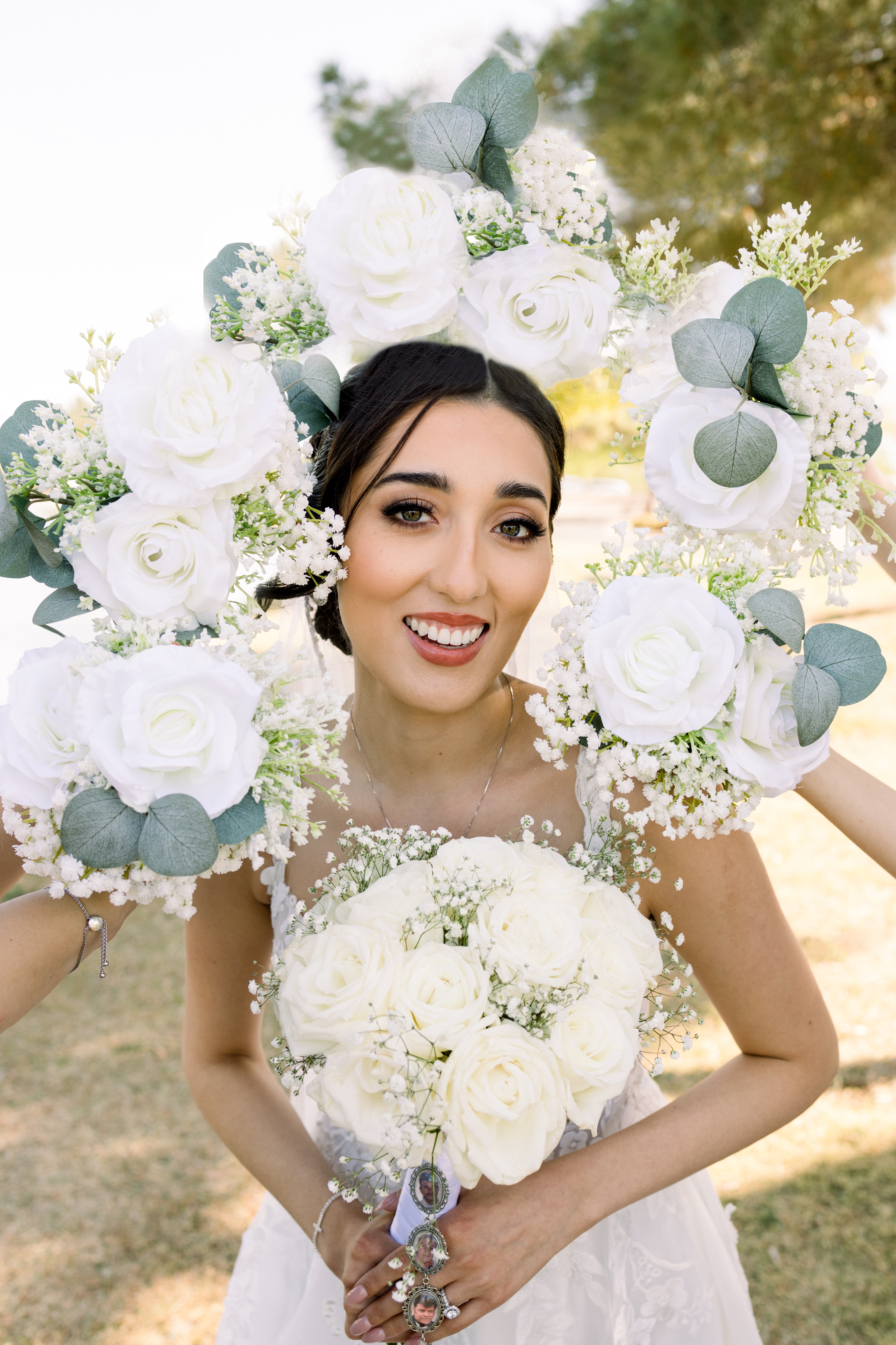 Smiling woman in wedding dress with floral headpiece and bouquet outdoors, trees in background.