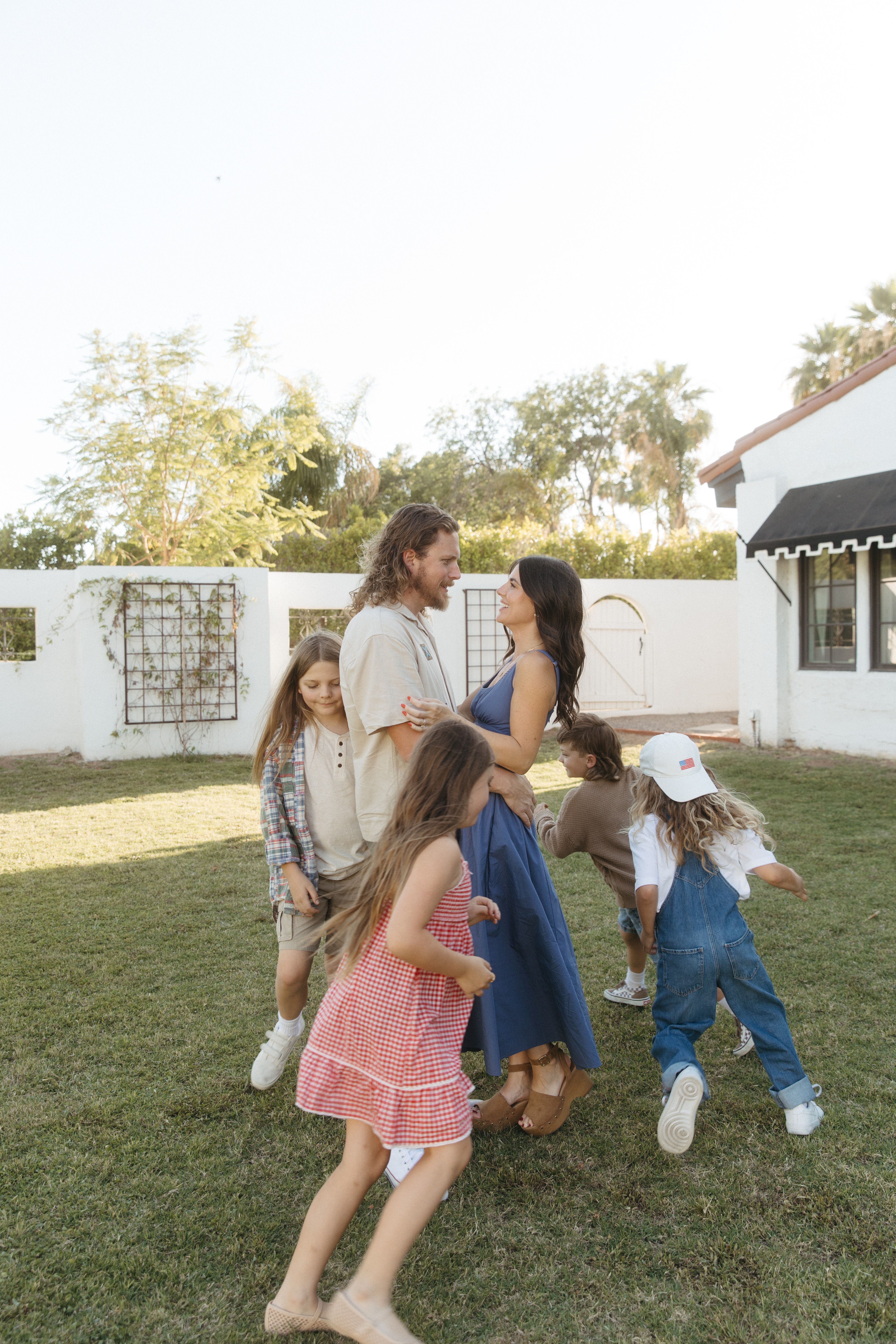 Group of children and a woman standing outdoors on grass, with trees and a building in the background.