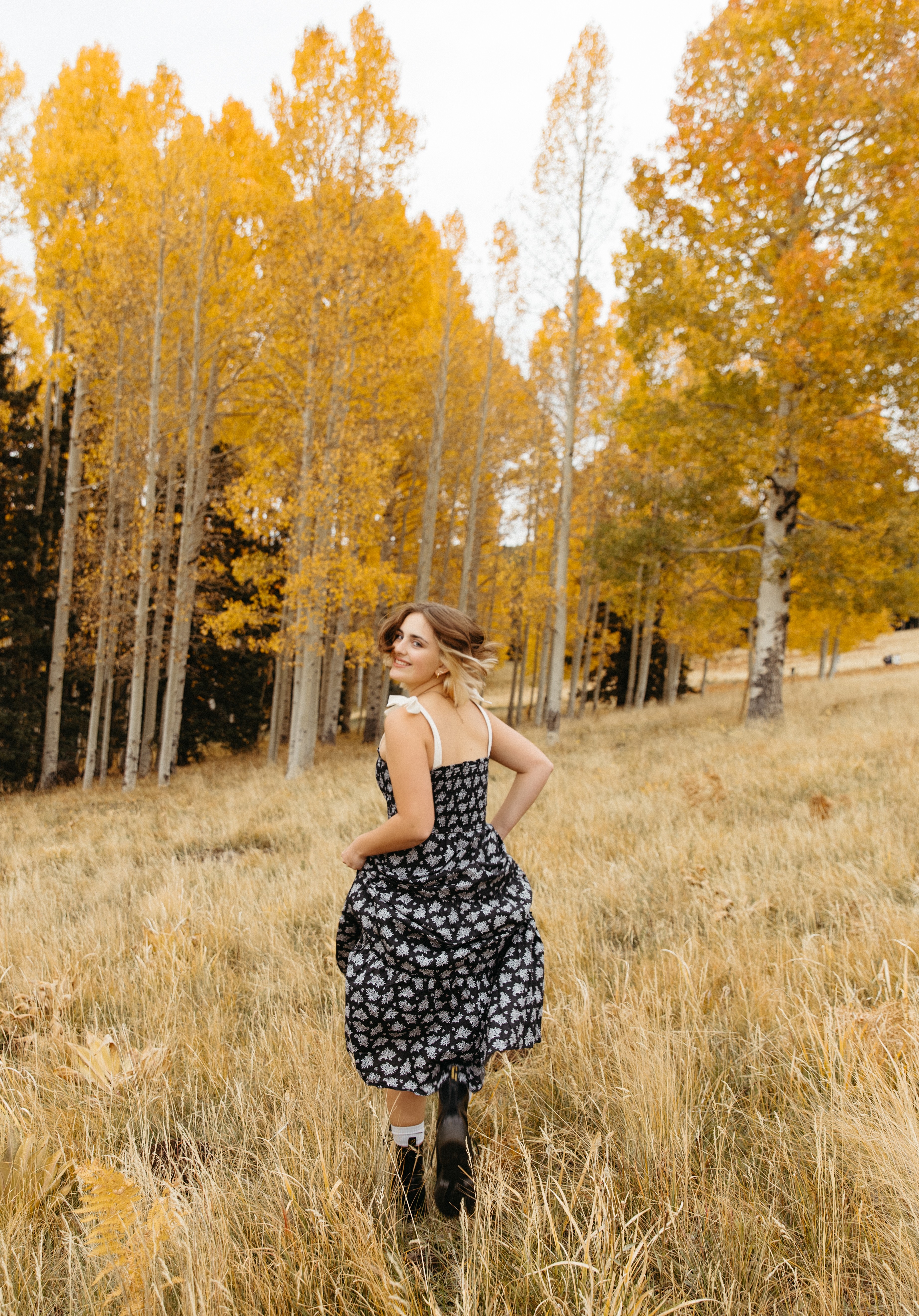 Woman in a black and white dress standing in a grassy field with yellow autumn trees in the background.