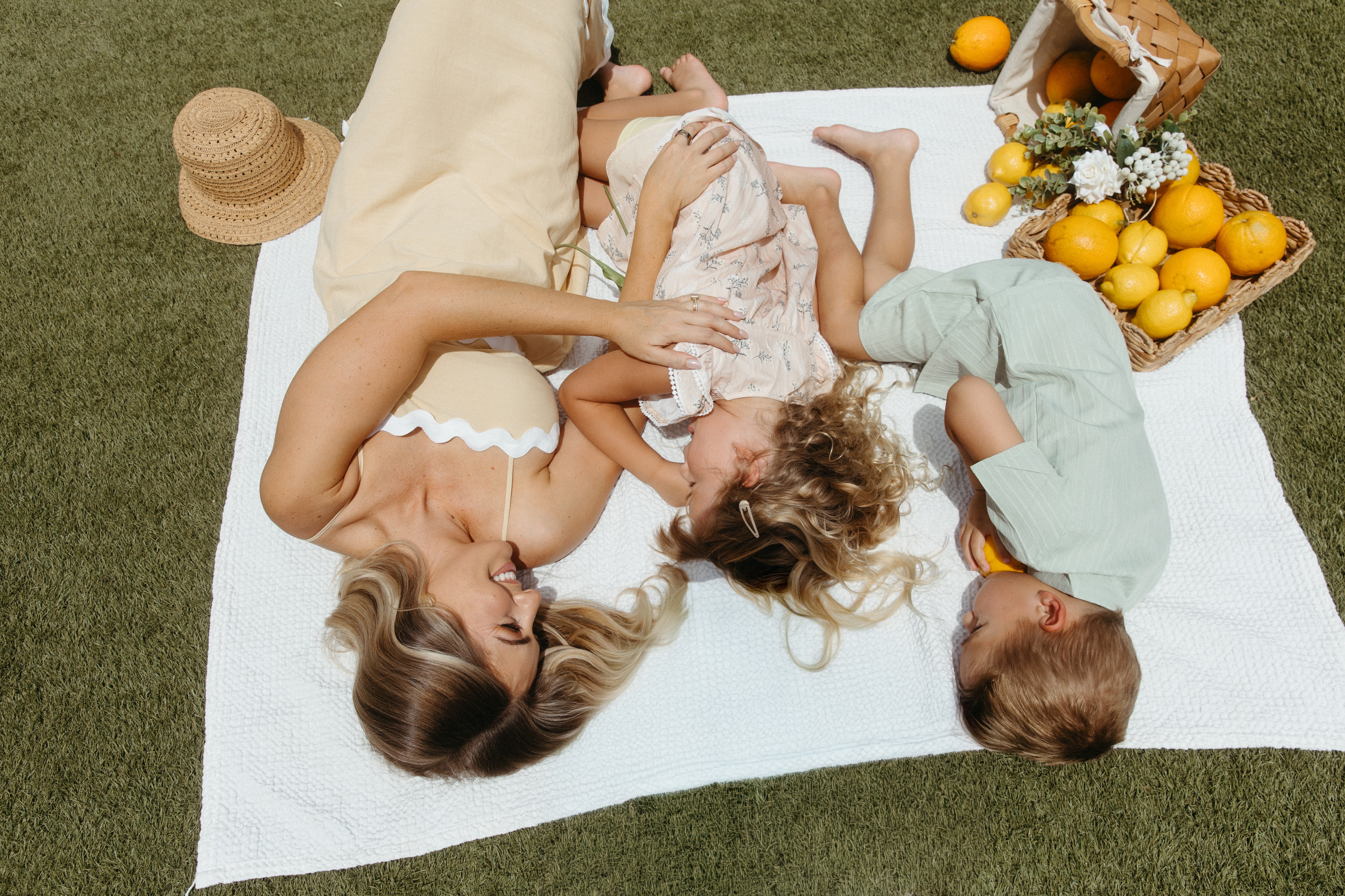 Woman, two children, and a dog lying on a white towel outdoors, with a basket of lemons and flowers nearby.