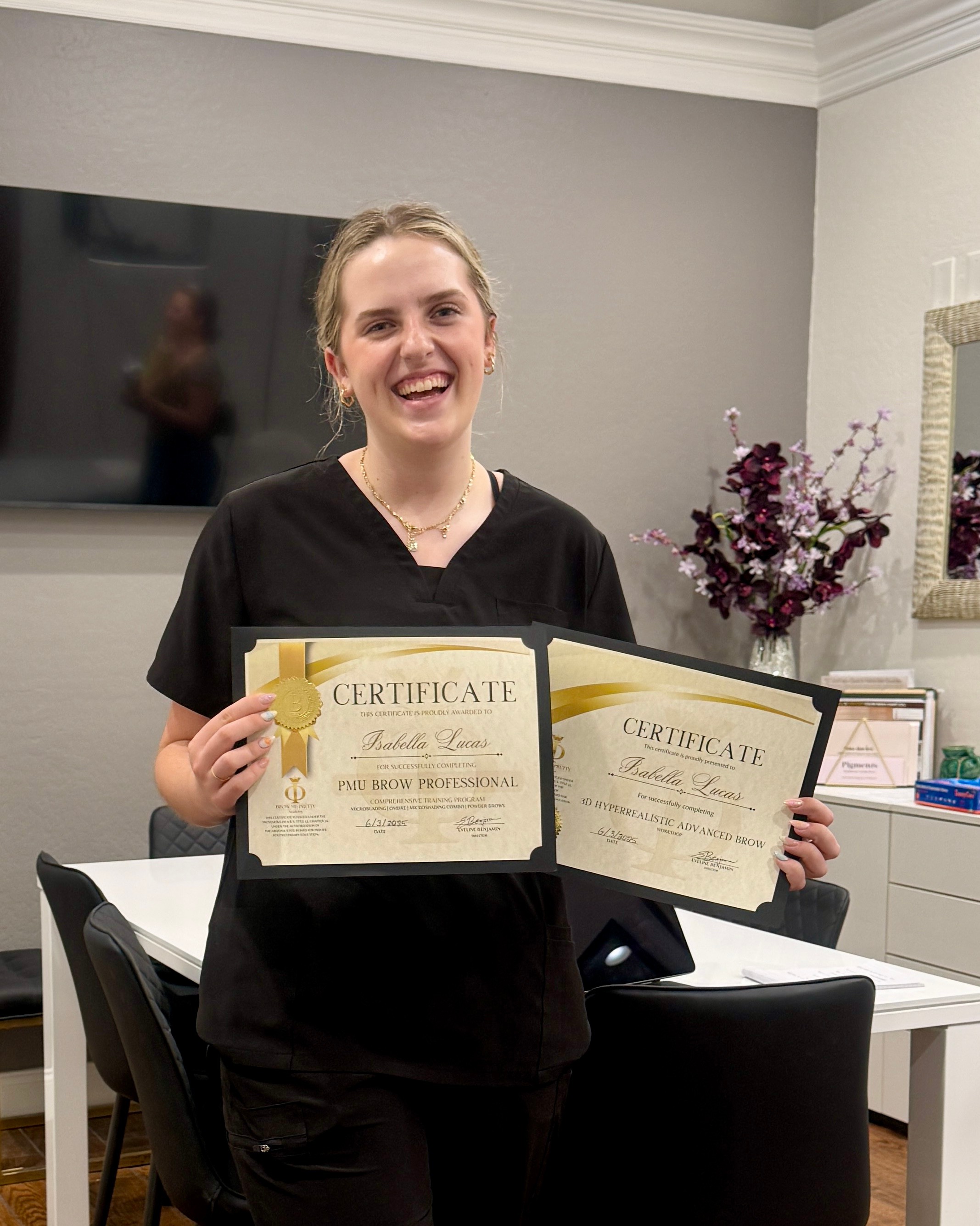 Young woman smiling, holding two certificates, standing in a room with a TV, flowers, and a desk.