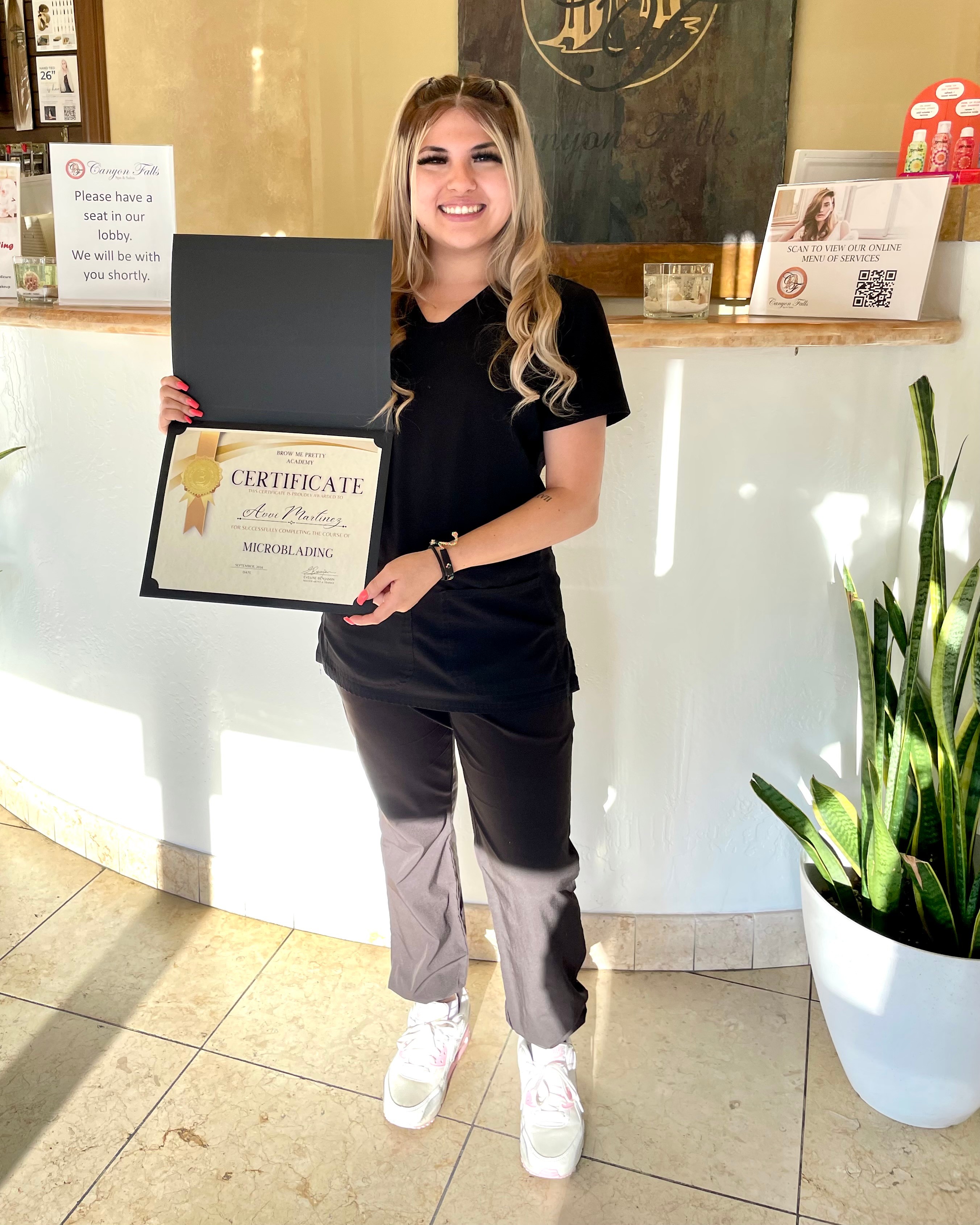 Smiling woman holding a certificate and a black folder in a reception area with a plant nearby.