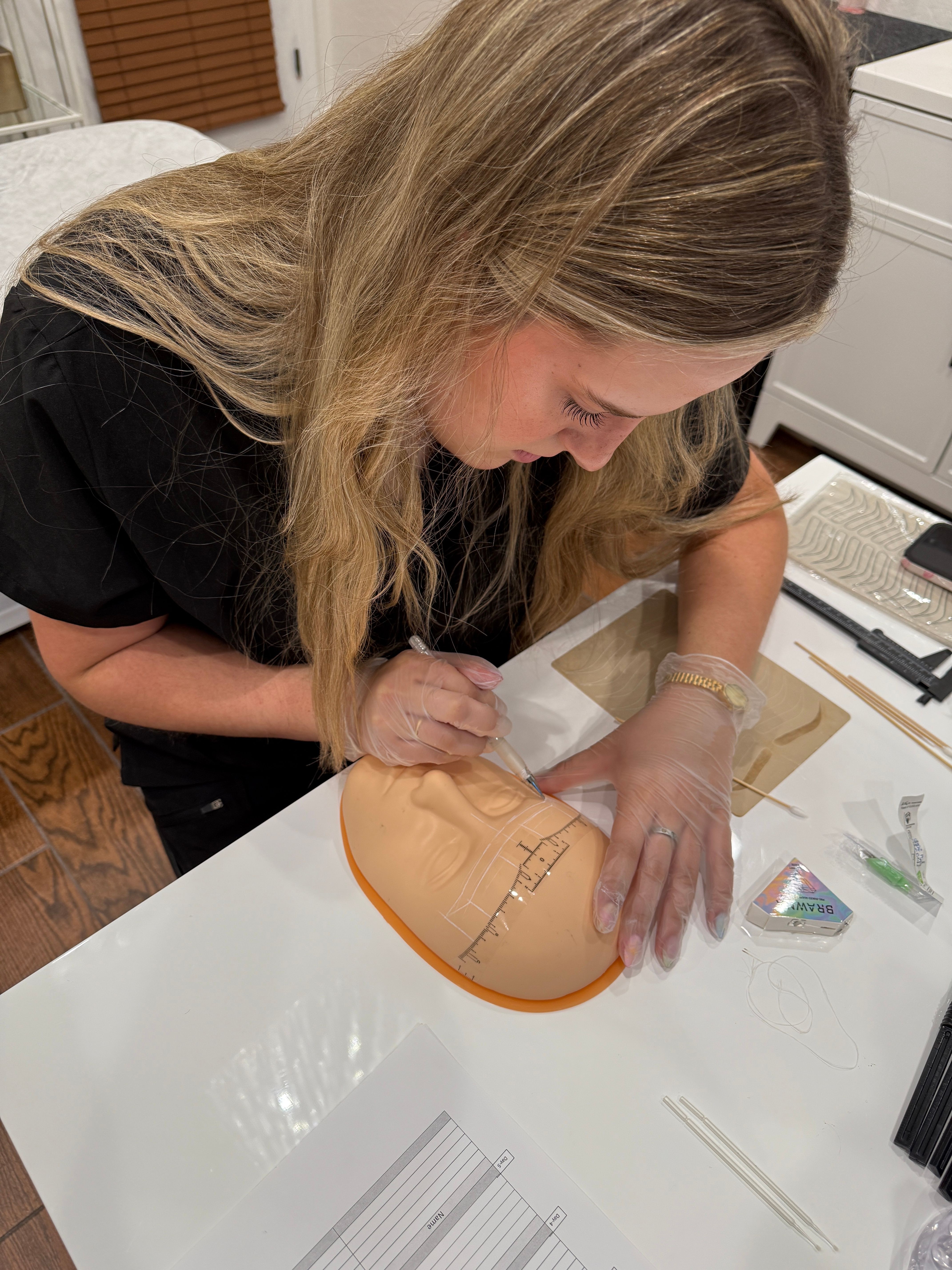 Woman with long blonde hair working on a craft project at a white table, using a tool on a circular object.