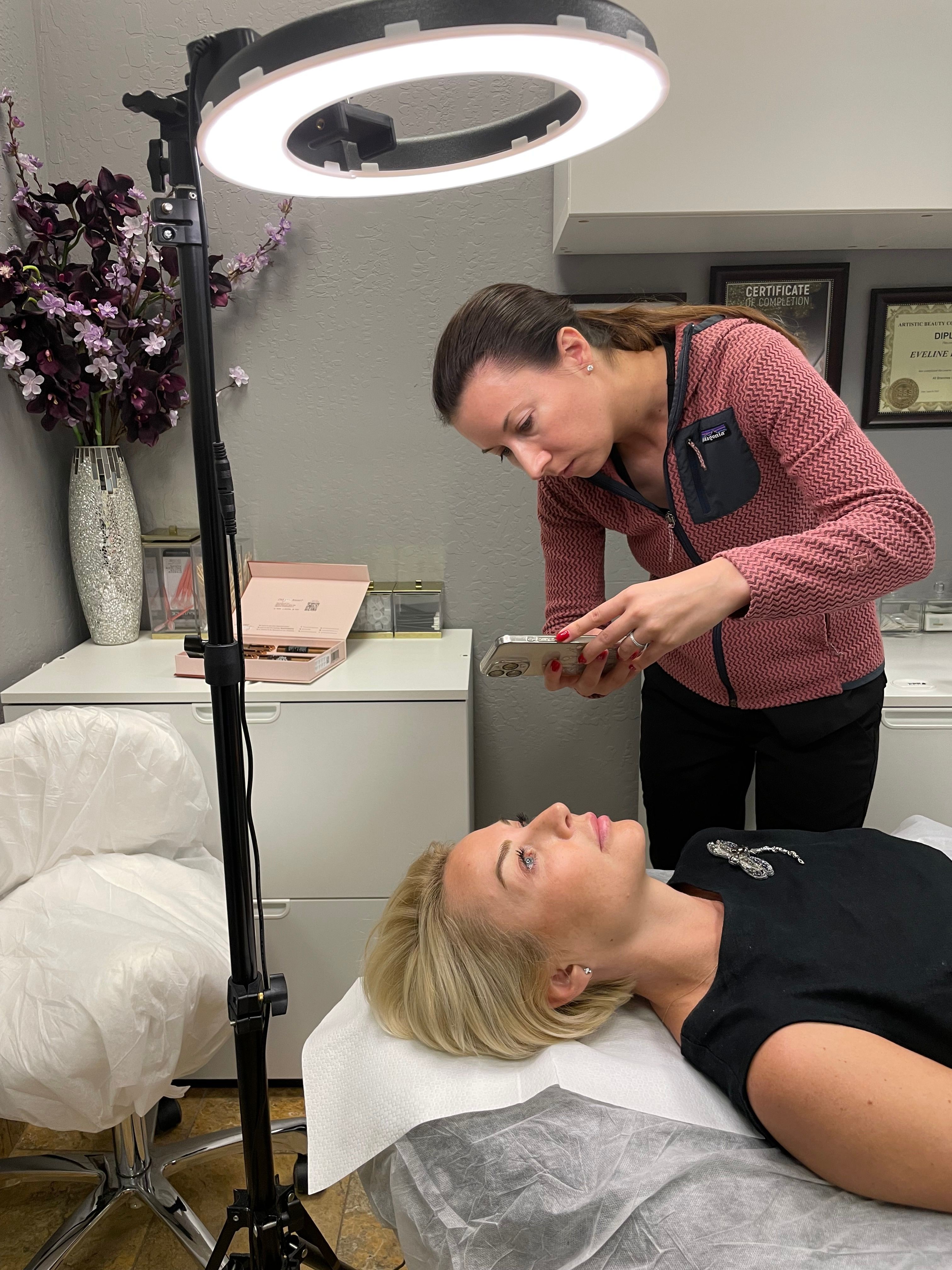 A woman is lying on a bed with her eyes closed, while a woman with a device near her face appears to be performing a procedure. The room has a white dresser, a pink box, and a flower arrangement.
