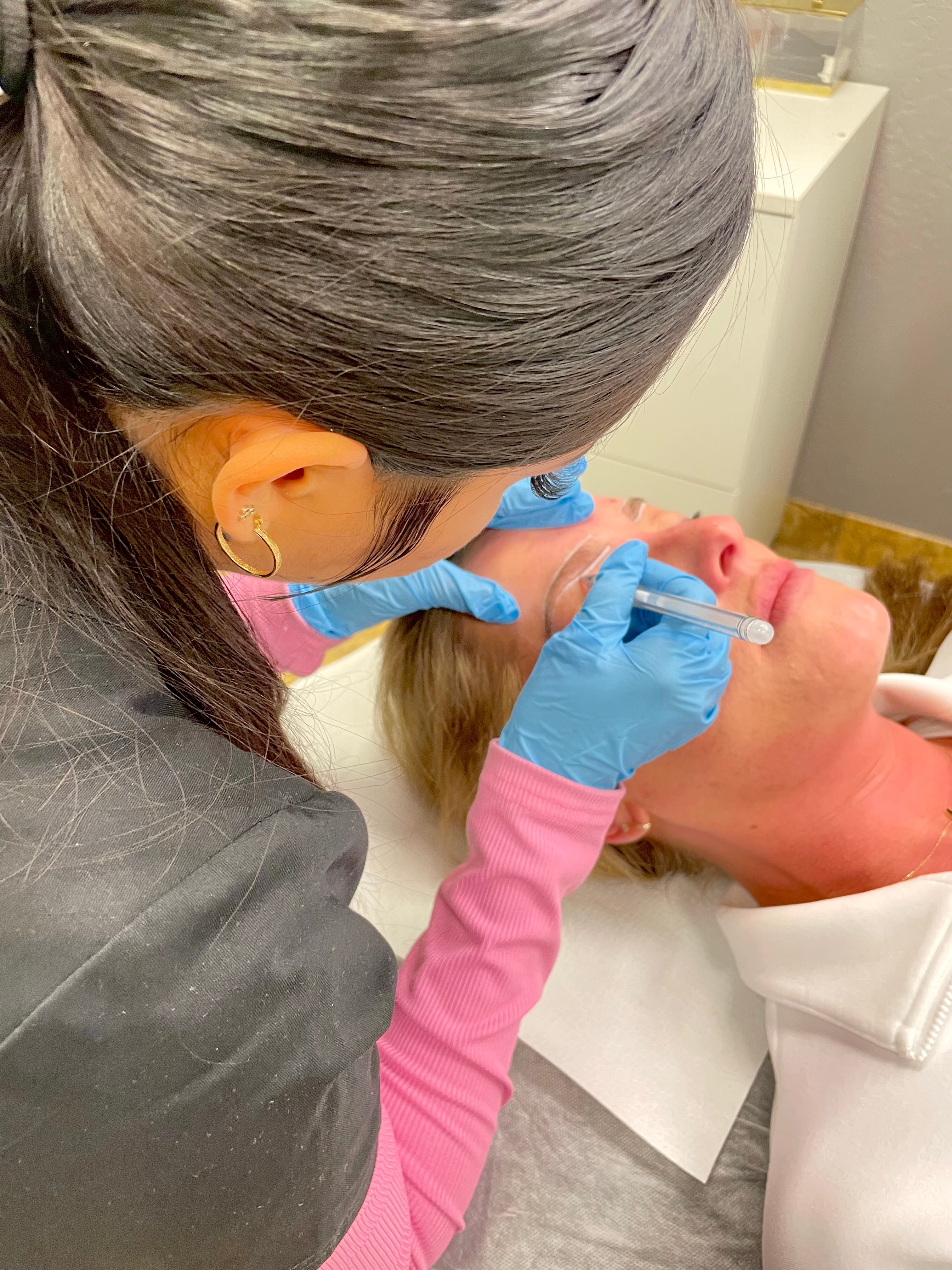 A person with gray hair and blue gloves is performing a cosmetic procedure on a woman lying down with her eyes closed.